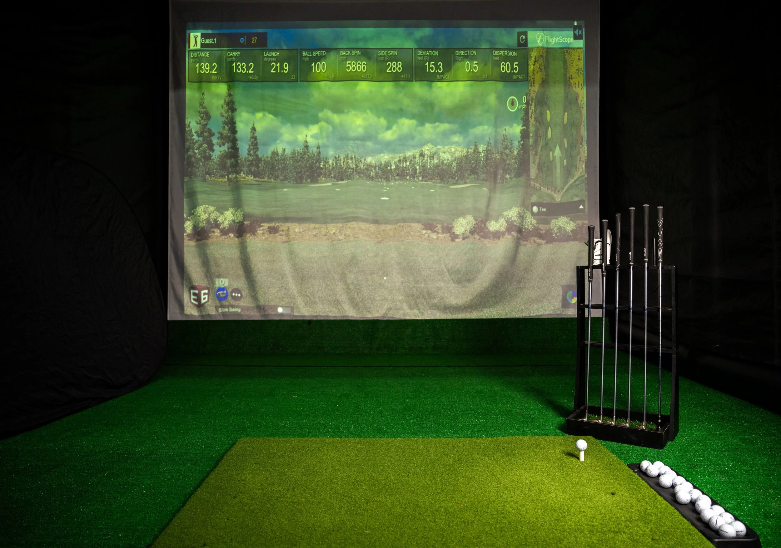 An indoor golf simulation room with a digital screen displaying a golf course, golf clubs in a stand, a golf tee with a ball on it, and a tray of golf balls on the floor.