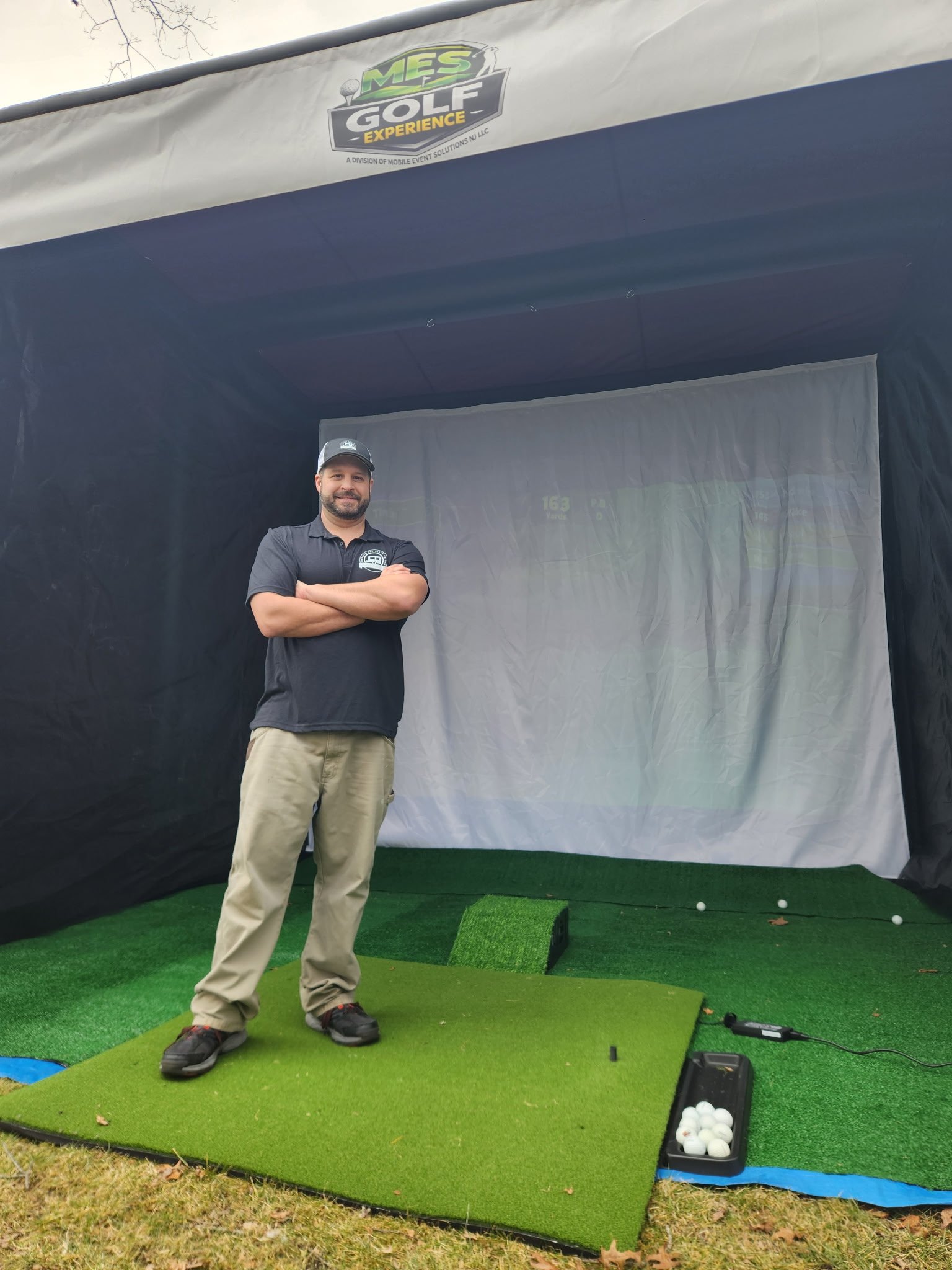 A man standing in a golf simulator studio with his arms crossed. The studio has a white projection screen, green artificial turf with golf balls, and a radar sensor device on the ground. The studio features a MES Golf Experience logo at the top.