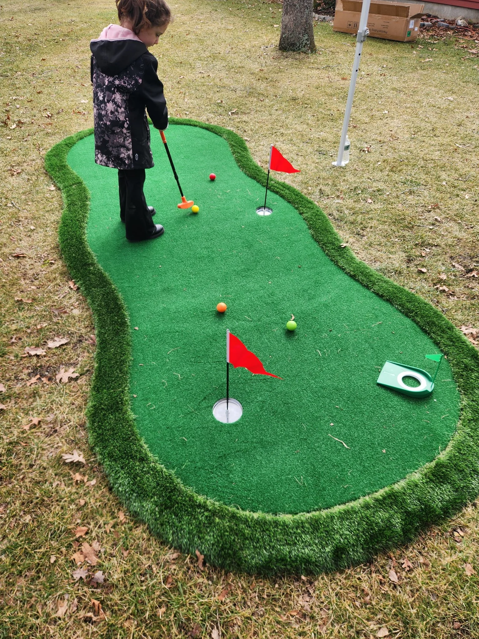 Child playing mini golf on a small putting green outdoors, with flags and a golf ball.