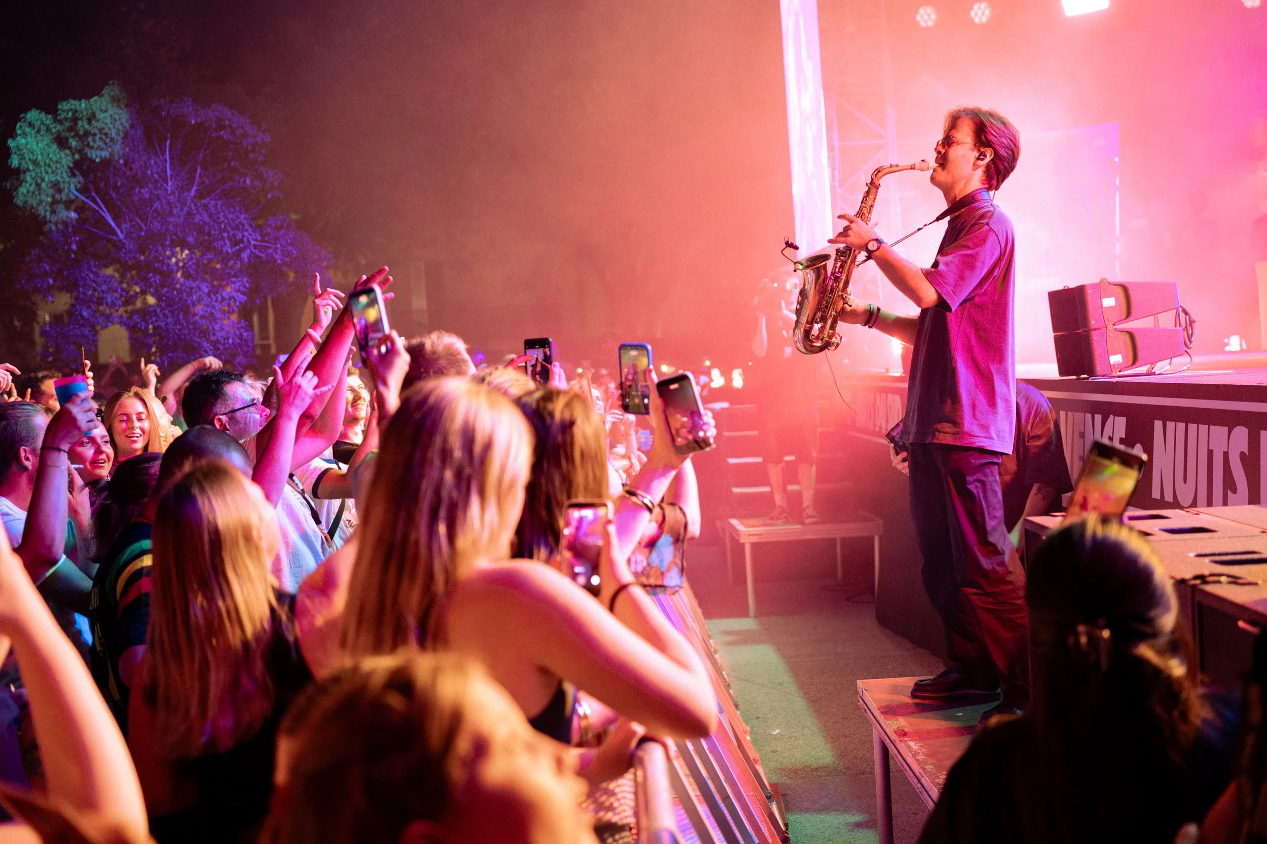 A lively concert scene with a crowd of young people taking photos and videos of a musician playing a saxophone on stage. The stage is bathed in colorful lights.