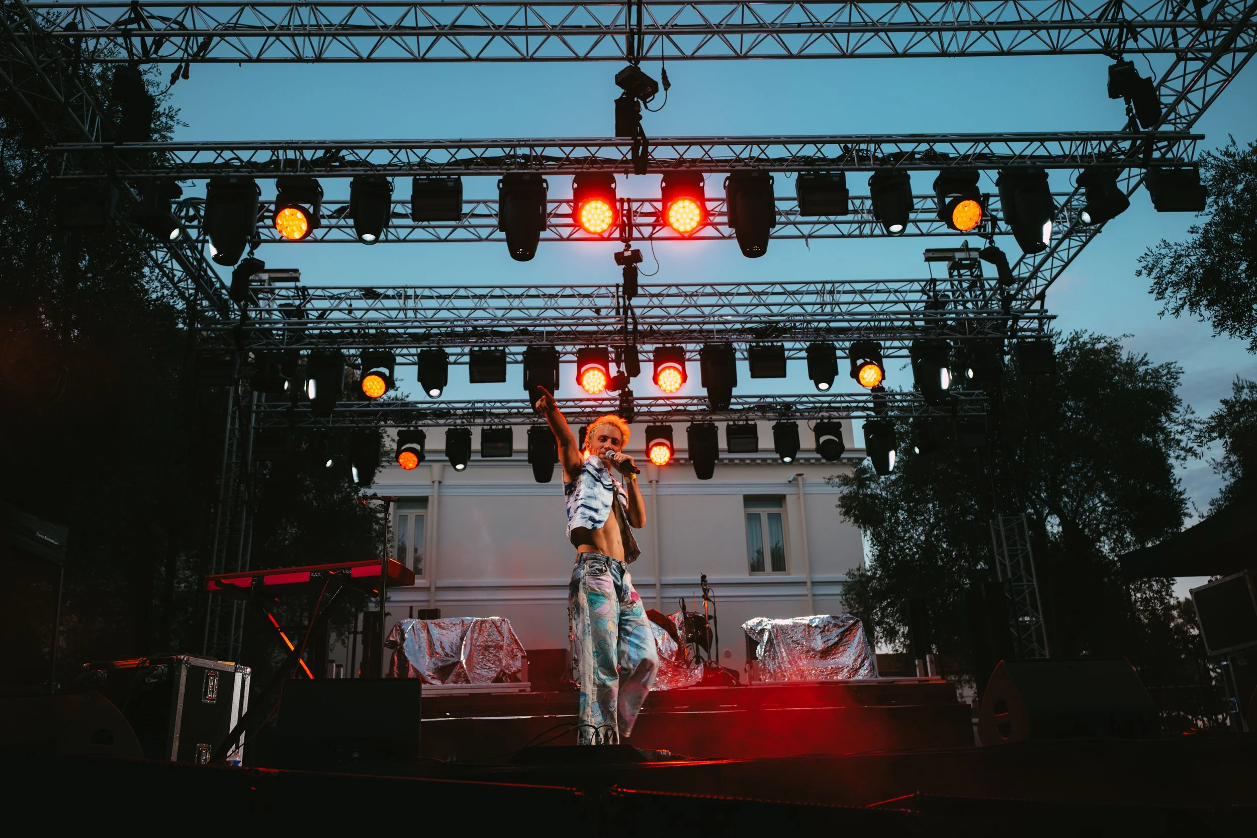 A performer singing into a microphone on an outdoor stage with red stage lights, metal truss structures, and a person in colorful clothing, during dusk with a clear sky and buildings in the background.