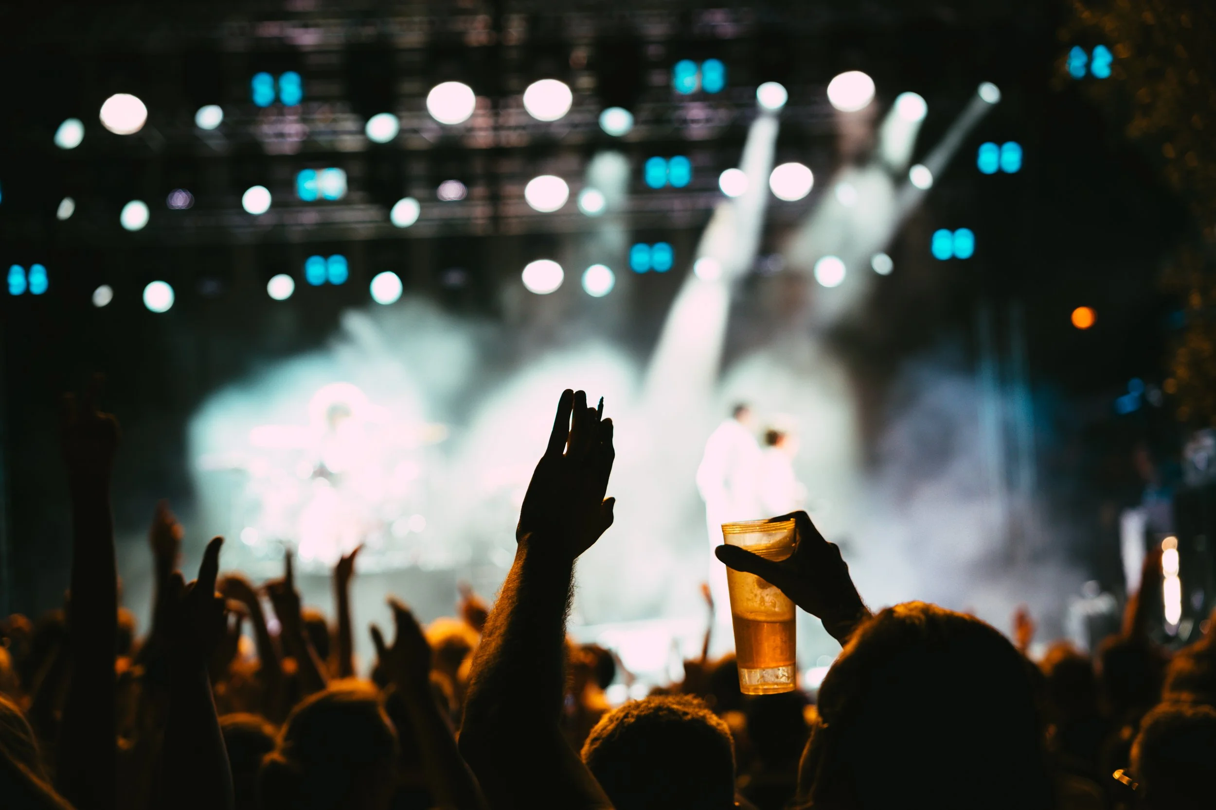 Concert crowd watching a stage with bright lights, with hands raised and one person holding a drink.