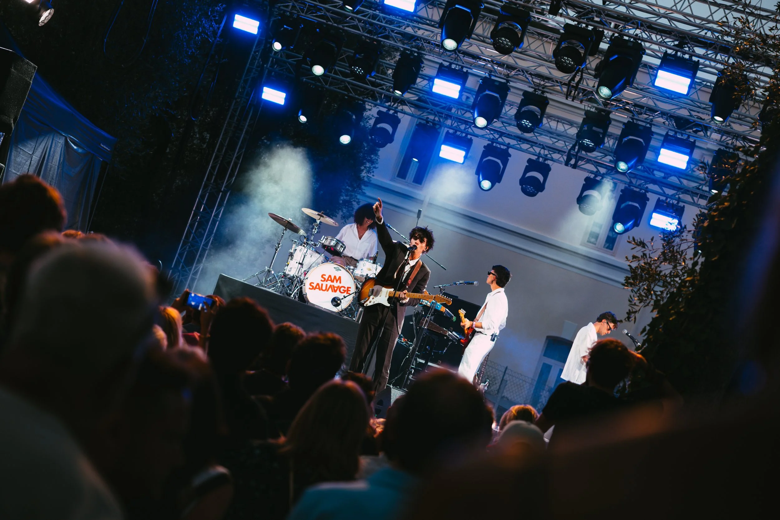 A band performs on an outdoor stage at night with blue and white lighting, row of musicians including a guitarist, drummer, and keyboard player, audience in the foreground.