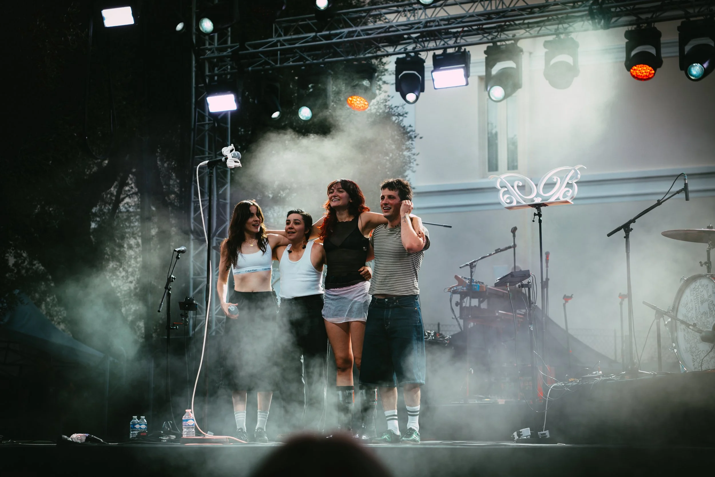 Four performers, three women and one man, standing together on an outdoor stage, smiling and embracing each other after a performance, with musical instruments and equipment visible in the background.