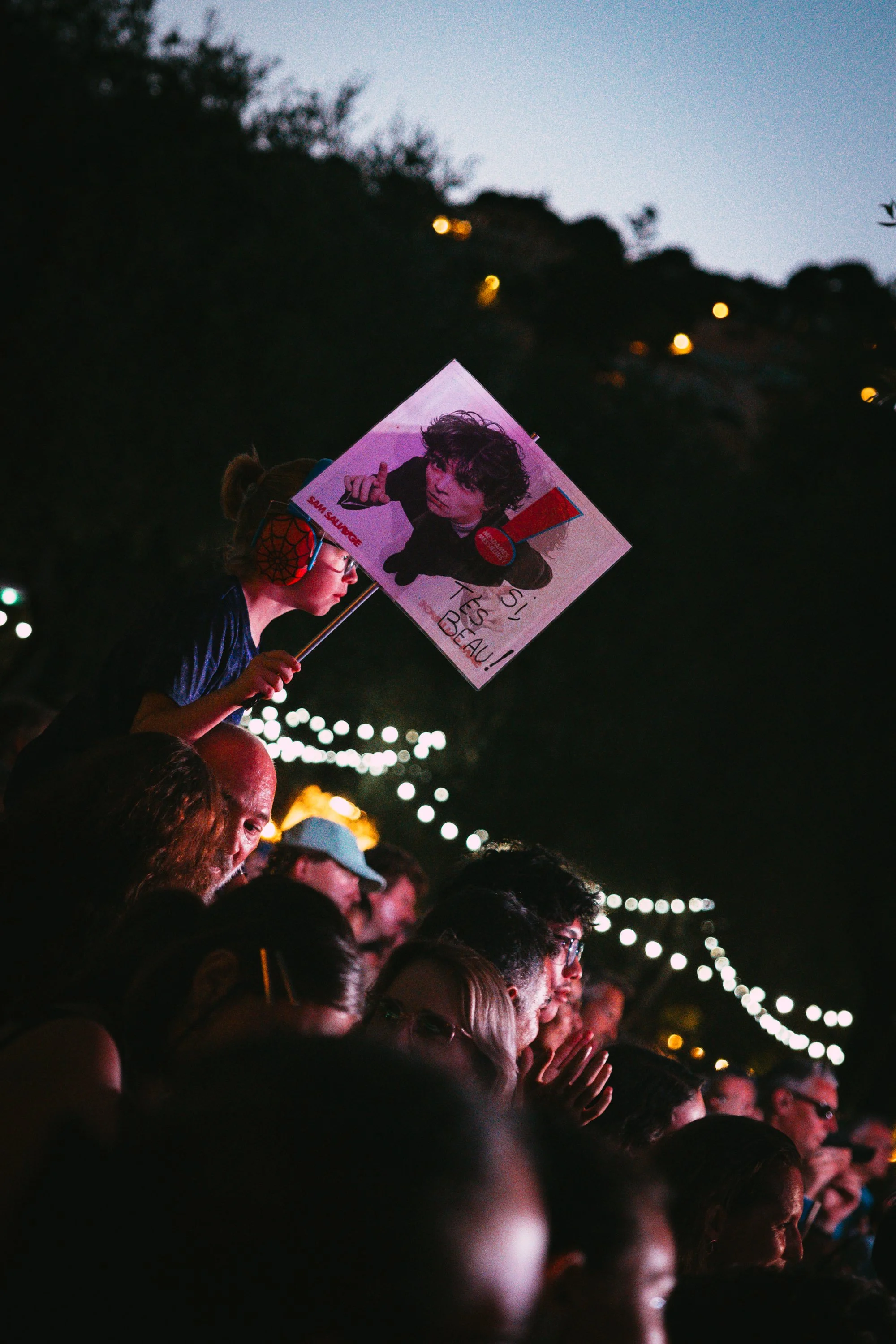 Crowd of people attending an outdoor event at dusk, with a woman holding a sign featuring a photo of a young man with curly hair, and using red and black protective headphones. String lights are visible above.