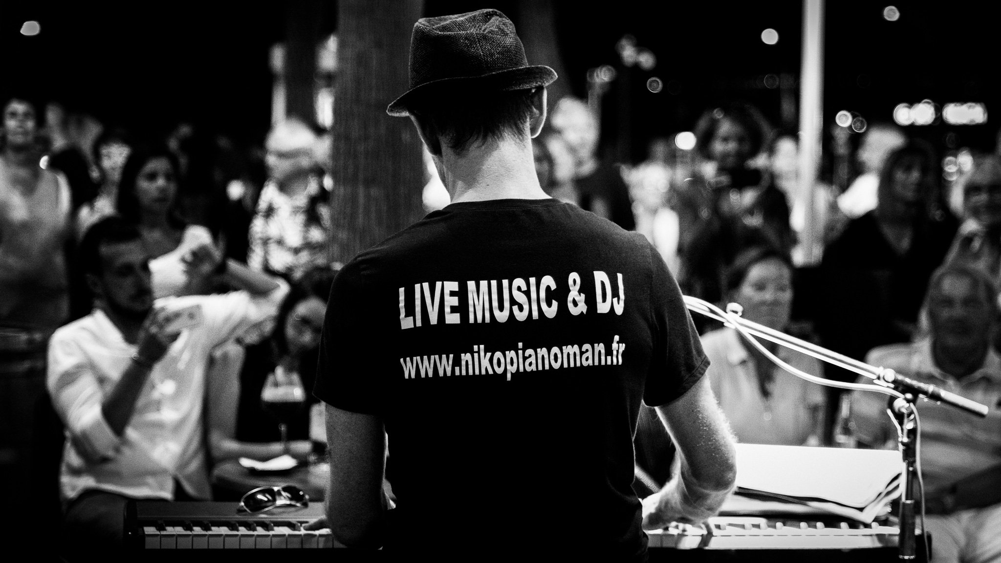 Back view of a musician wearing a hat and a shirt with the words 'LIVE MUSIC & DJ' and a website, performing on stage with a keyboard while audience members watch.