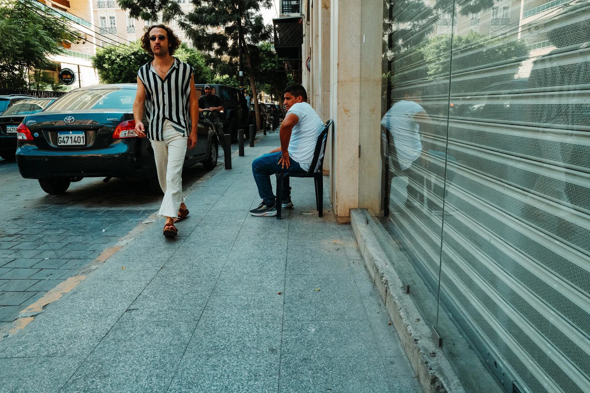 A man walking on the sidewalk wearing a black-and-white striped sleeveless shirt, white pants, and sunglasses. Another man in a white shirt and blue jeans sitting on a chair against a building. Parked cars and a row of trees line the street in the ba