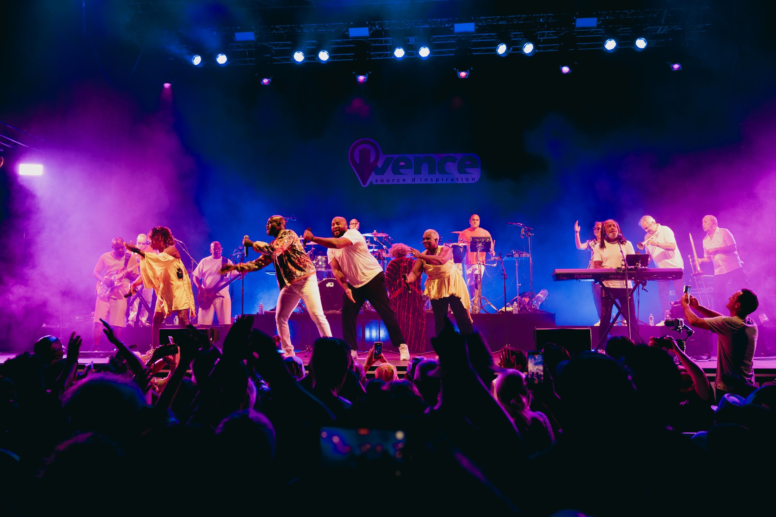 A live musical performance on stage with multiple singers and musicians, colorful stage lighting including purple and blue hues, a crowd in front taking photos, and a sign that reads 'Vence, source d'inspiration' in the background.