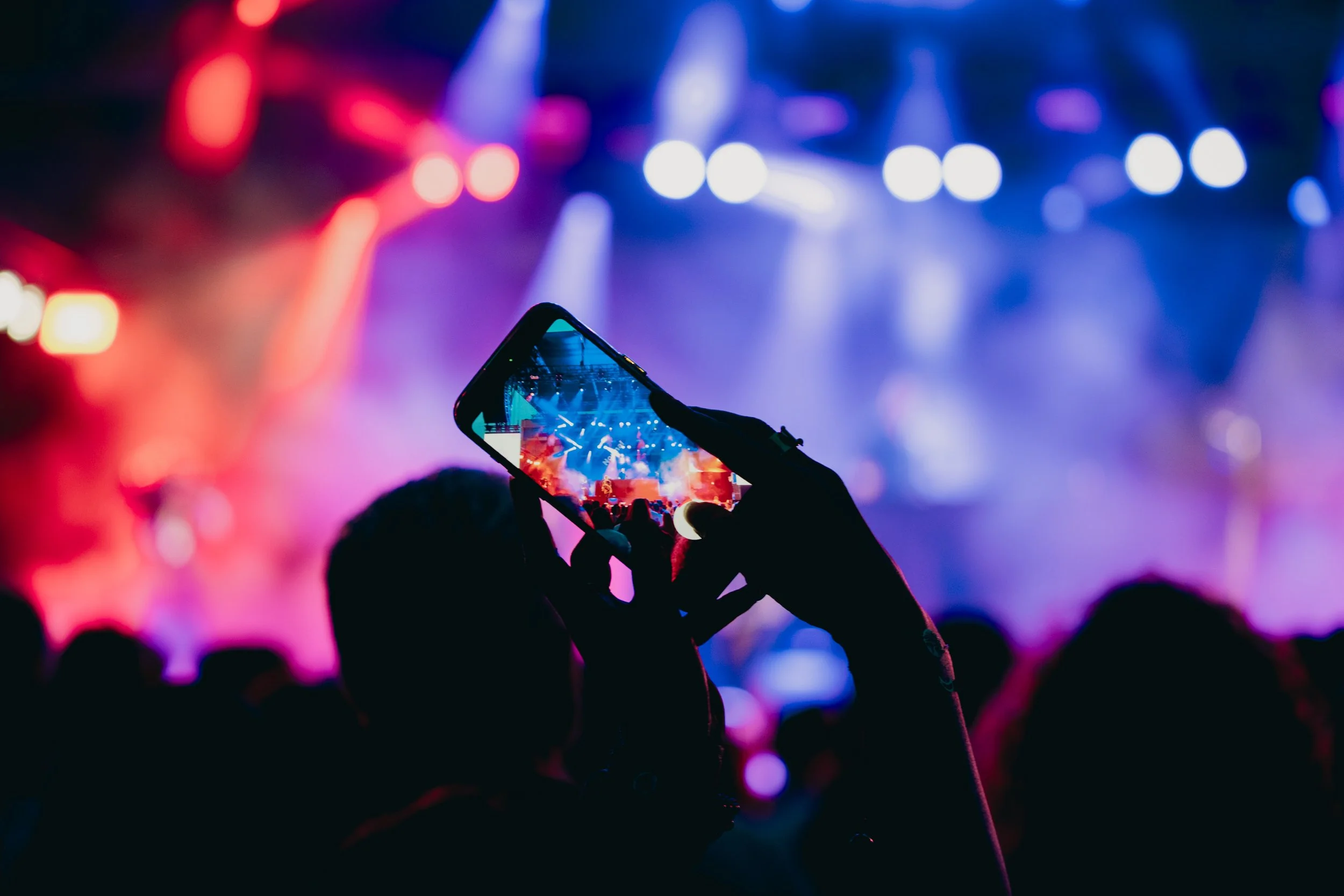 Person holding up a smartphone capturing a concert or stage performance with colorful lights in the background.