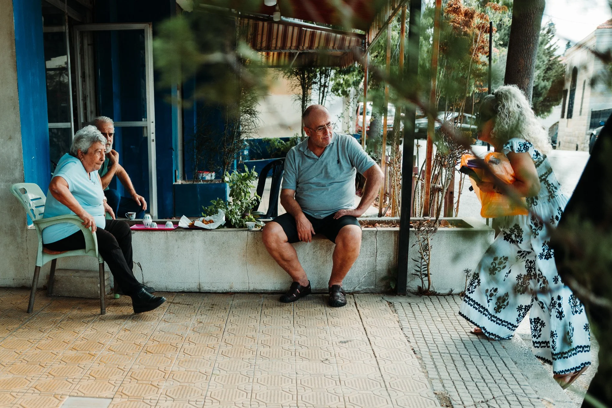 A woman with curly blonde hair wearing a white and blue patterned dress is talking to three elderly people seated outside a building. The elderly woman is on a white plastic chair, and the elderly man is behind her, on a concrete window ledge. The ma
