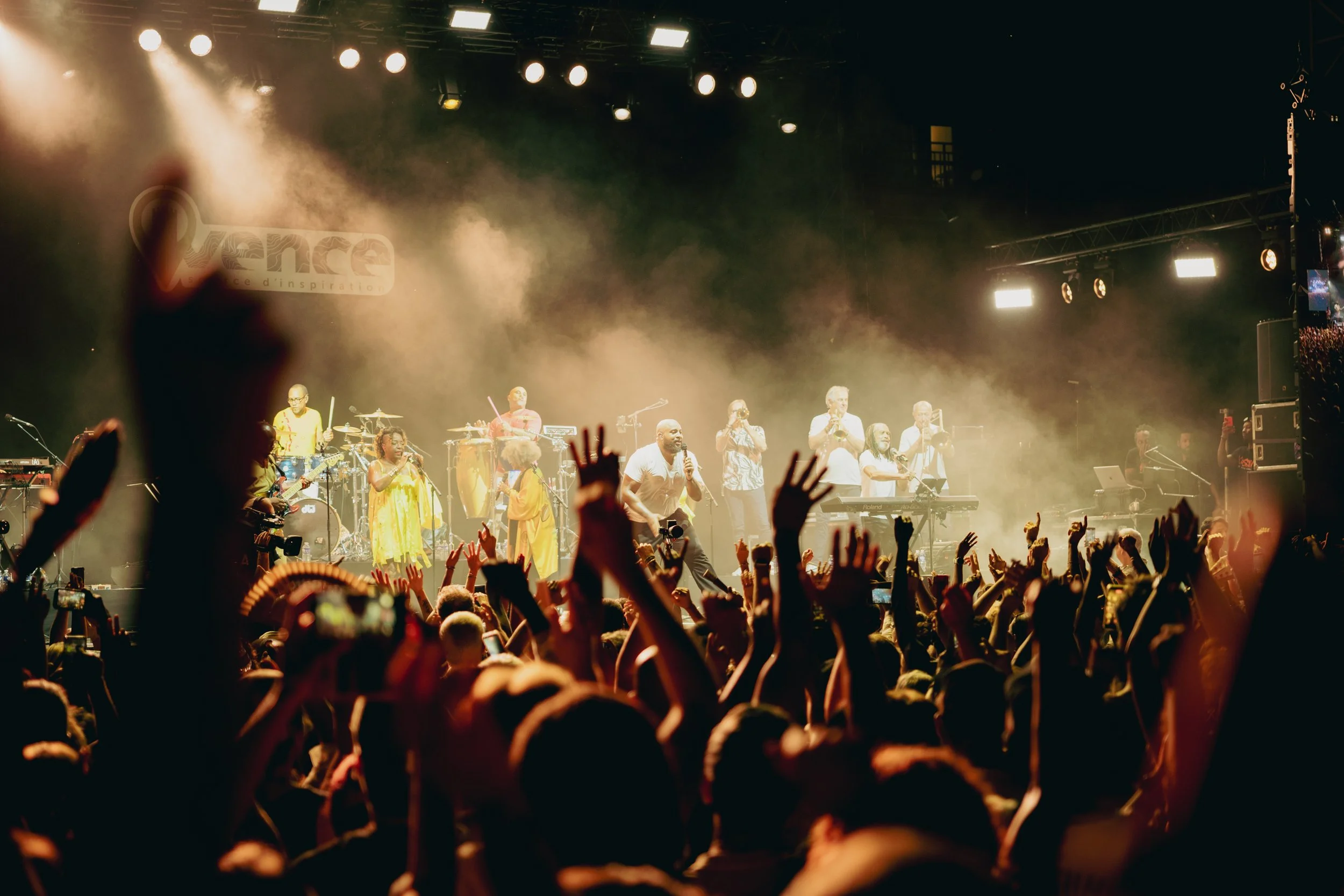 Crowd at a concert with music band on stage under bright stage lights, hands raised in the air.