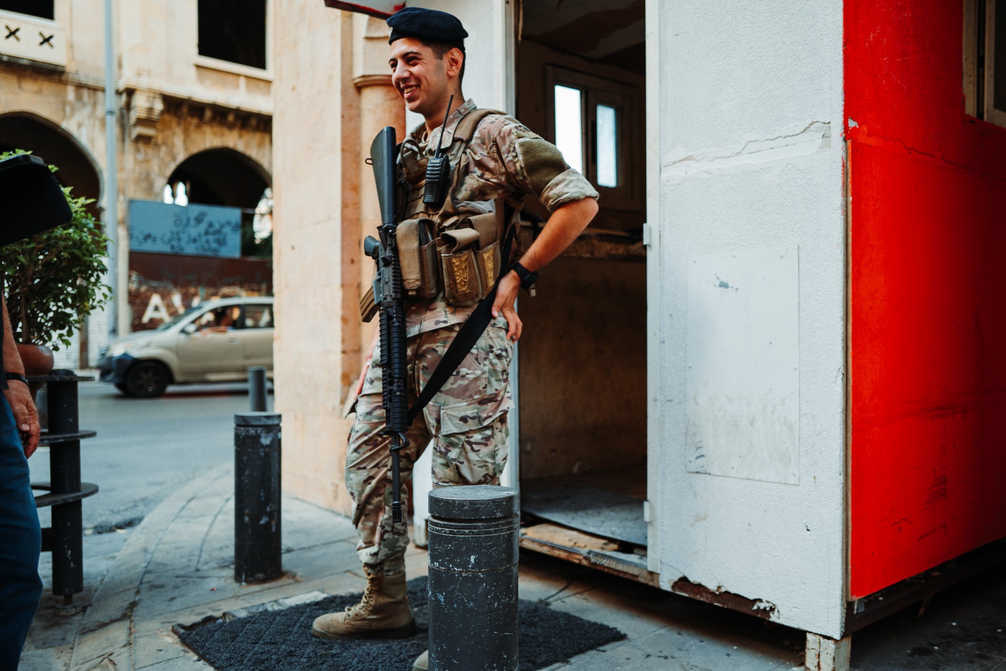 A smiling soldier in camouflage uniform and black beret standing outside a building, holding a rifle, next to a small structure on a city sidewalk.