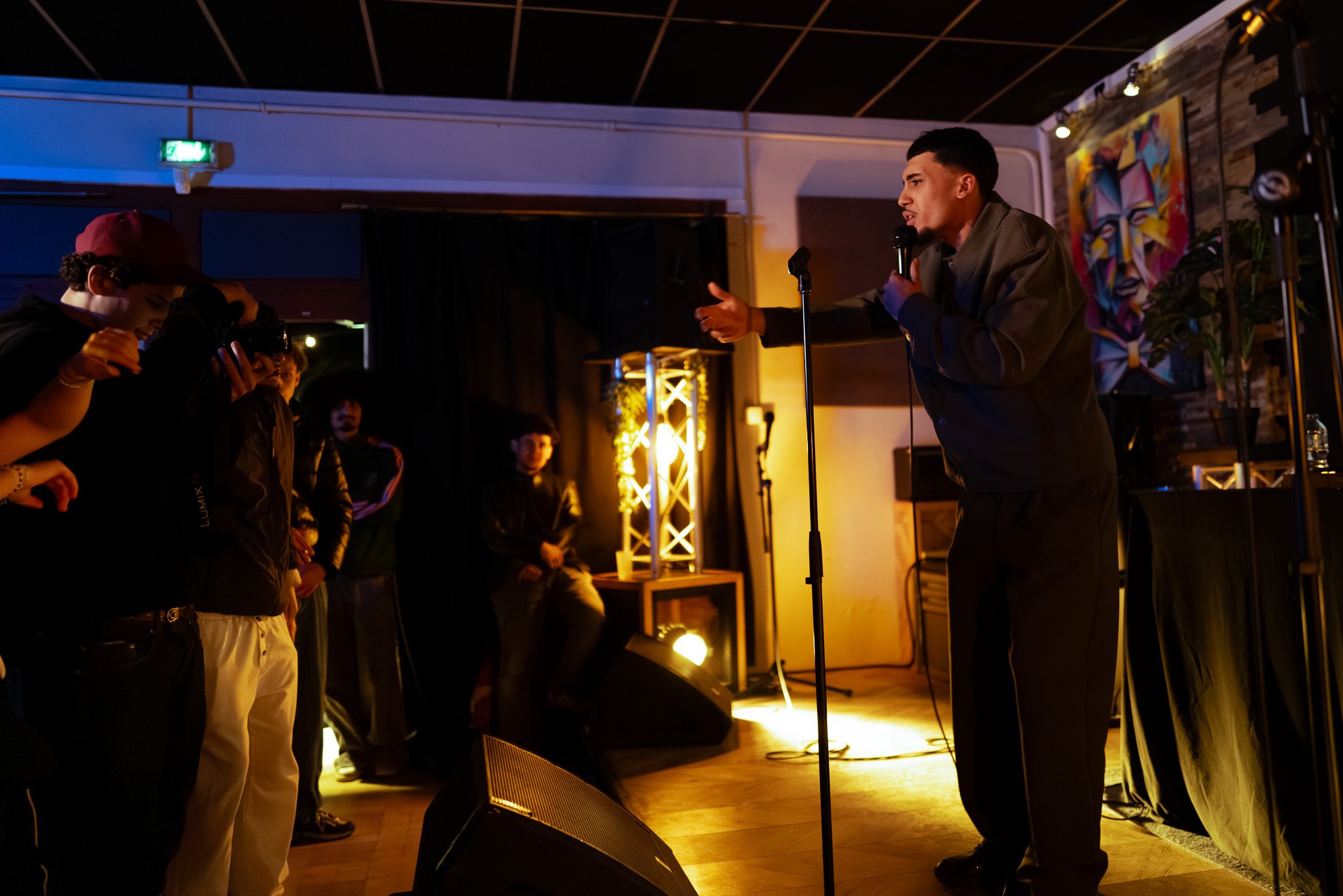 A man performing stand-up comedy on stage with a microphone, audience members watching, and stage lighting in a cozy indoor venue.