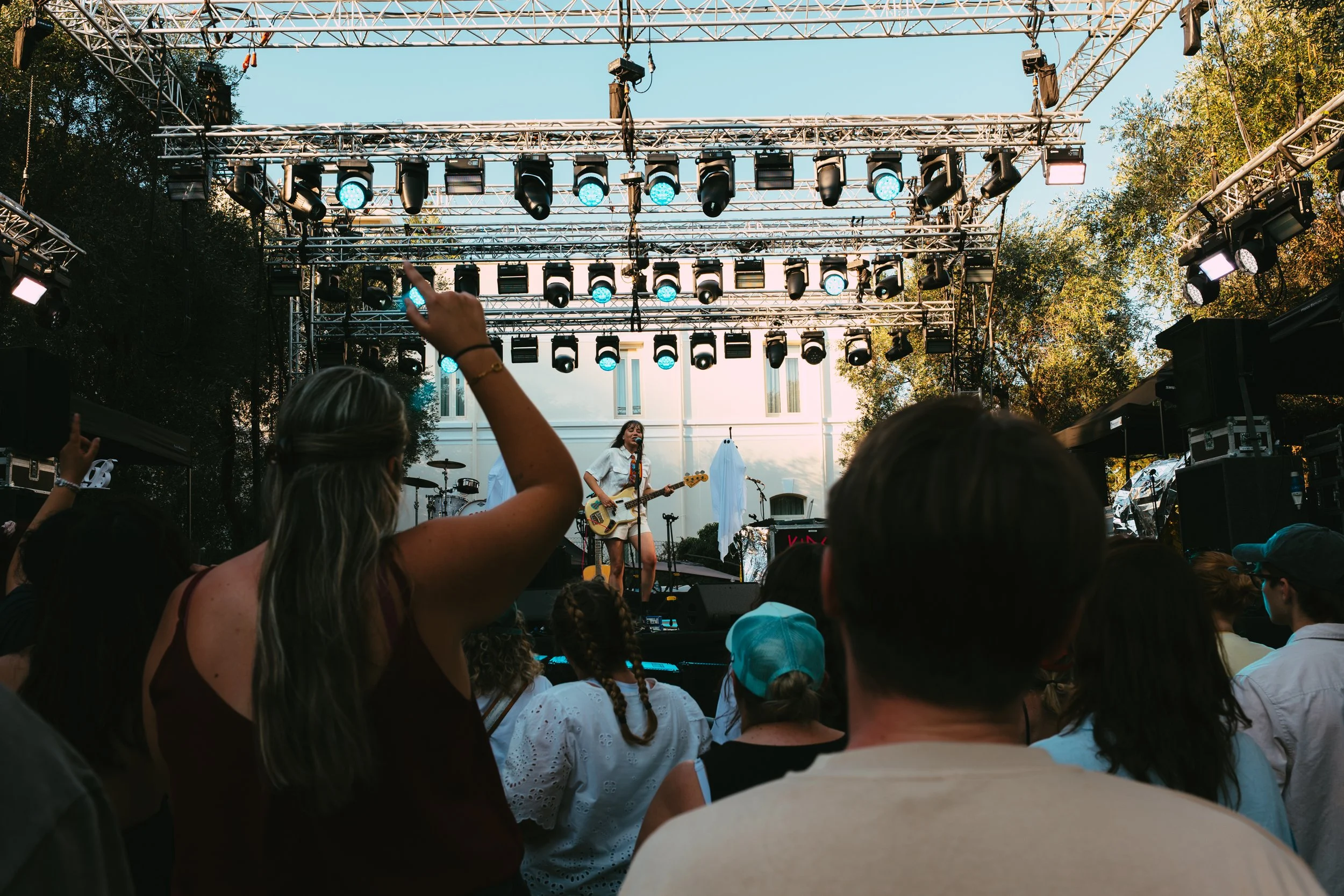 An outdoor concert stage with a performer playing guitar and singing, surrounded by crowd members under a clear sky with trees in the background.