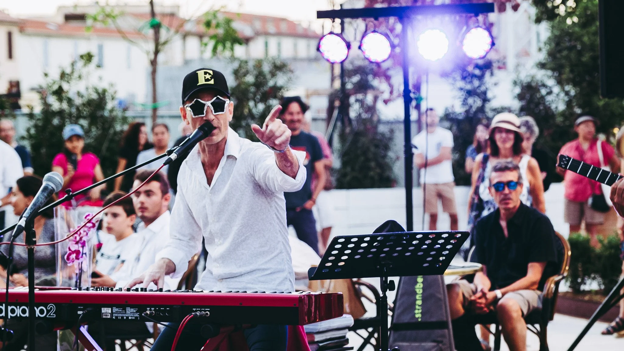 A man wearing sunglasses with star-shaped glasses and a black cap performing at an outdoor event, playing a keyboard and singing into a microphone, with an audience watching.