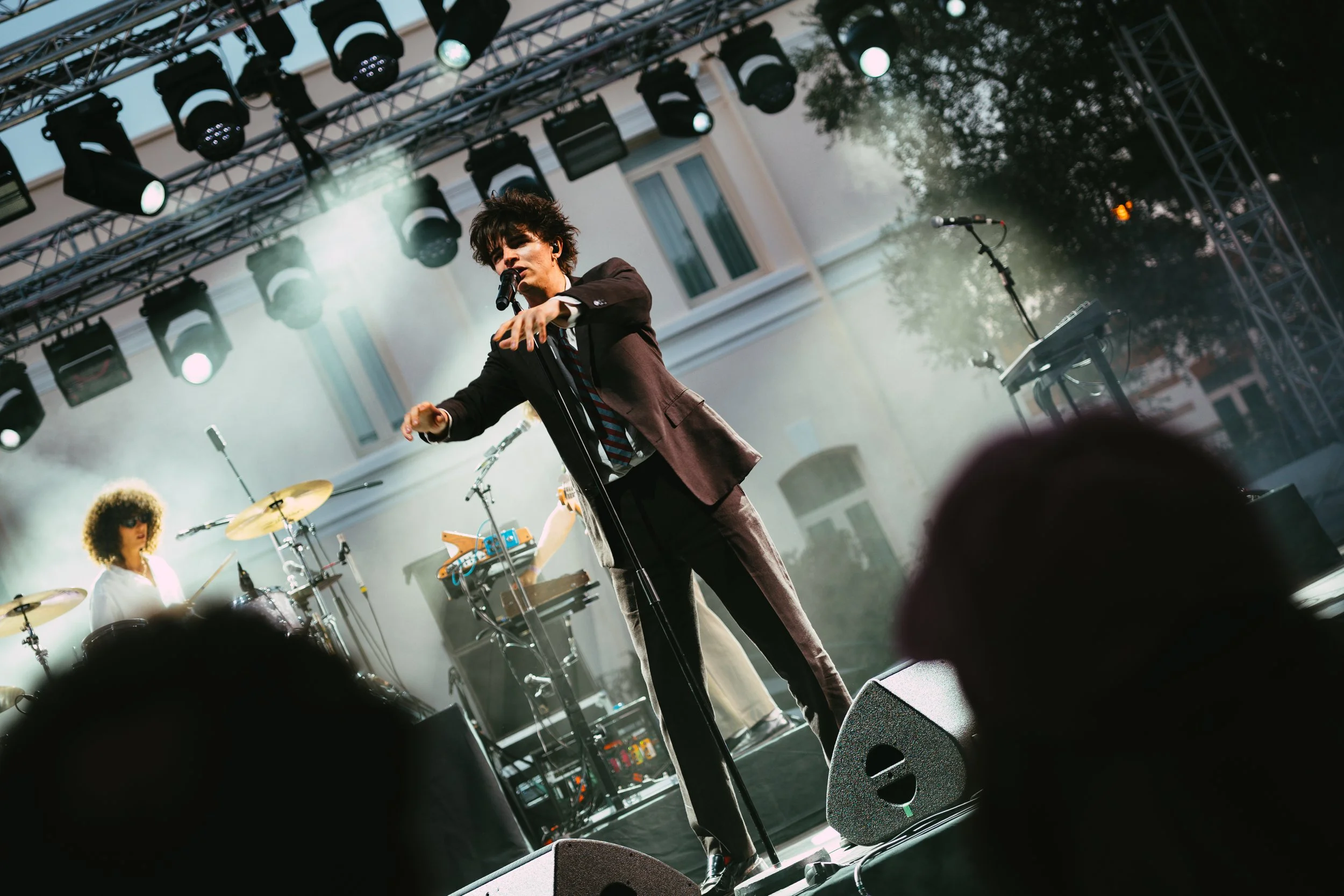 A singer performing on an outdoor stage at sunset, with a drummer and music equipment behind him, stage lights above, and audience heads in the foreground.