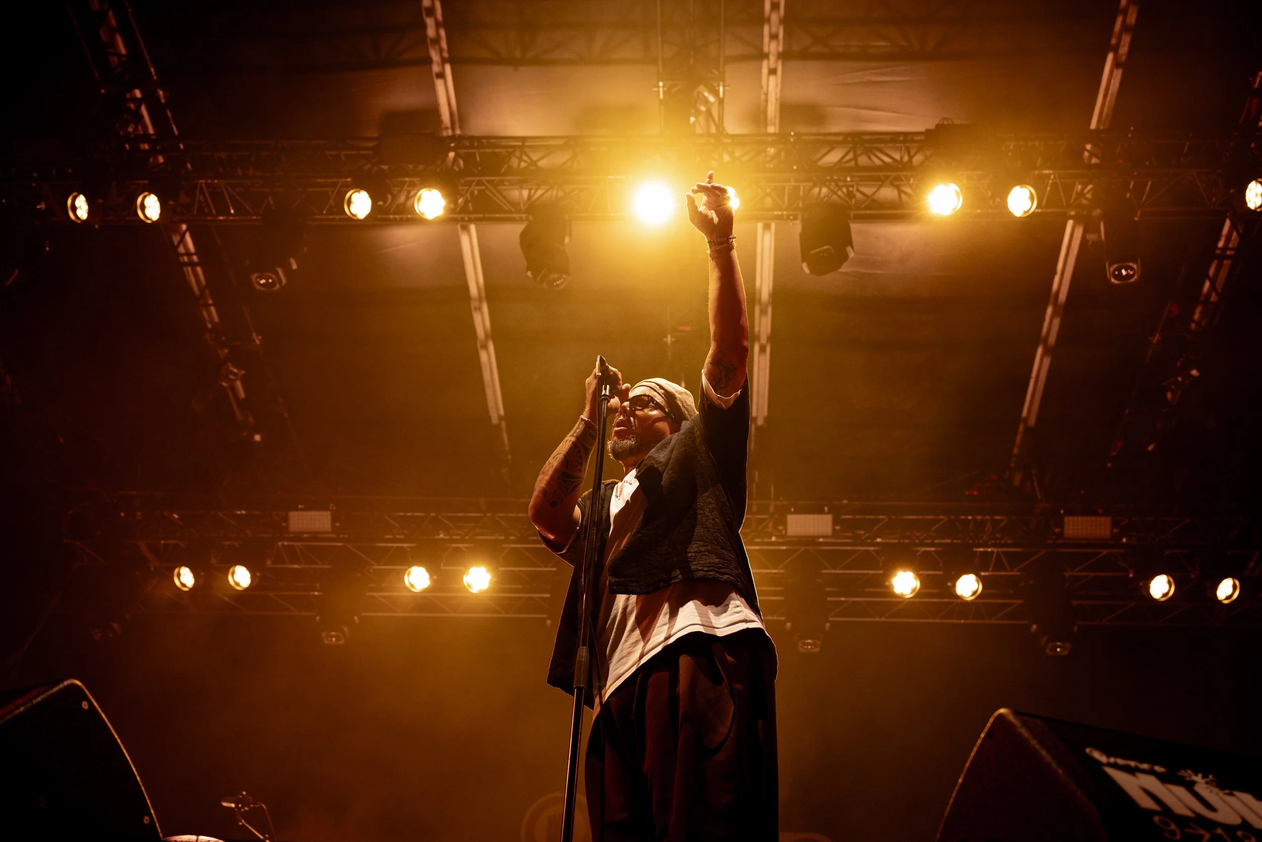 A musician performing on stage under bright yellowish-orange stage lights. The musician is holding a microphone with one hand and raising the other hand with a gesture, wearing glasses, a head wrap, and tattoos are visible on the arms.
