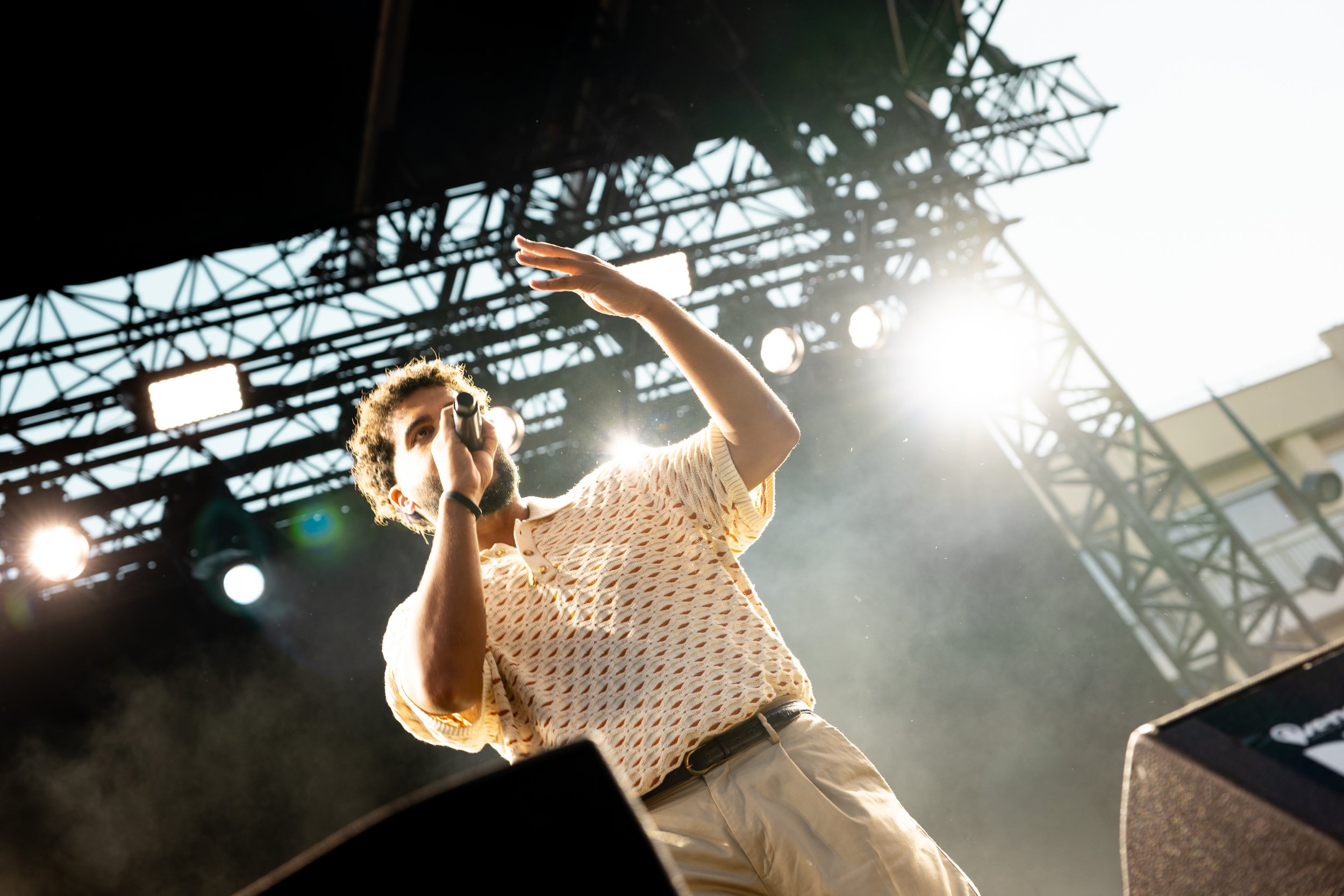 A male performer with curly hair and a beard singing into a microphone on stage, reaching out with his hand, with bright stage lights and scaffolding in the background.