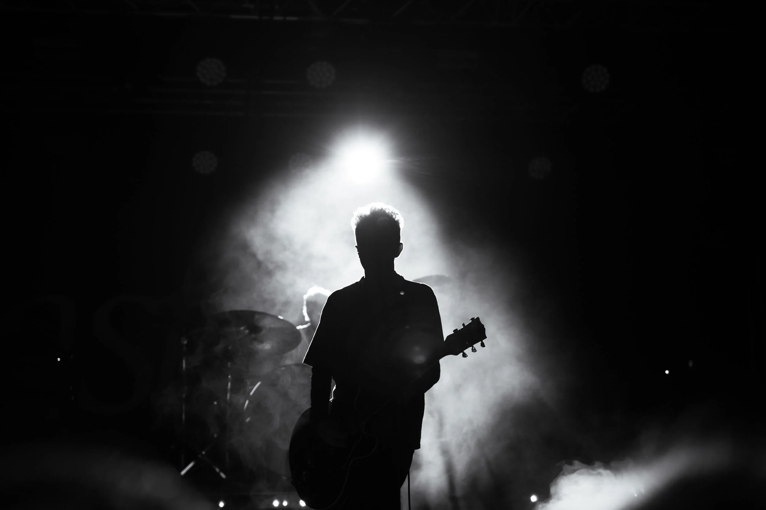 Silhouette of a musician playing guitar on stage, backlit by bright stage lights creating a dramatic effect.