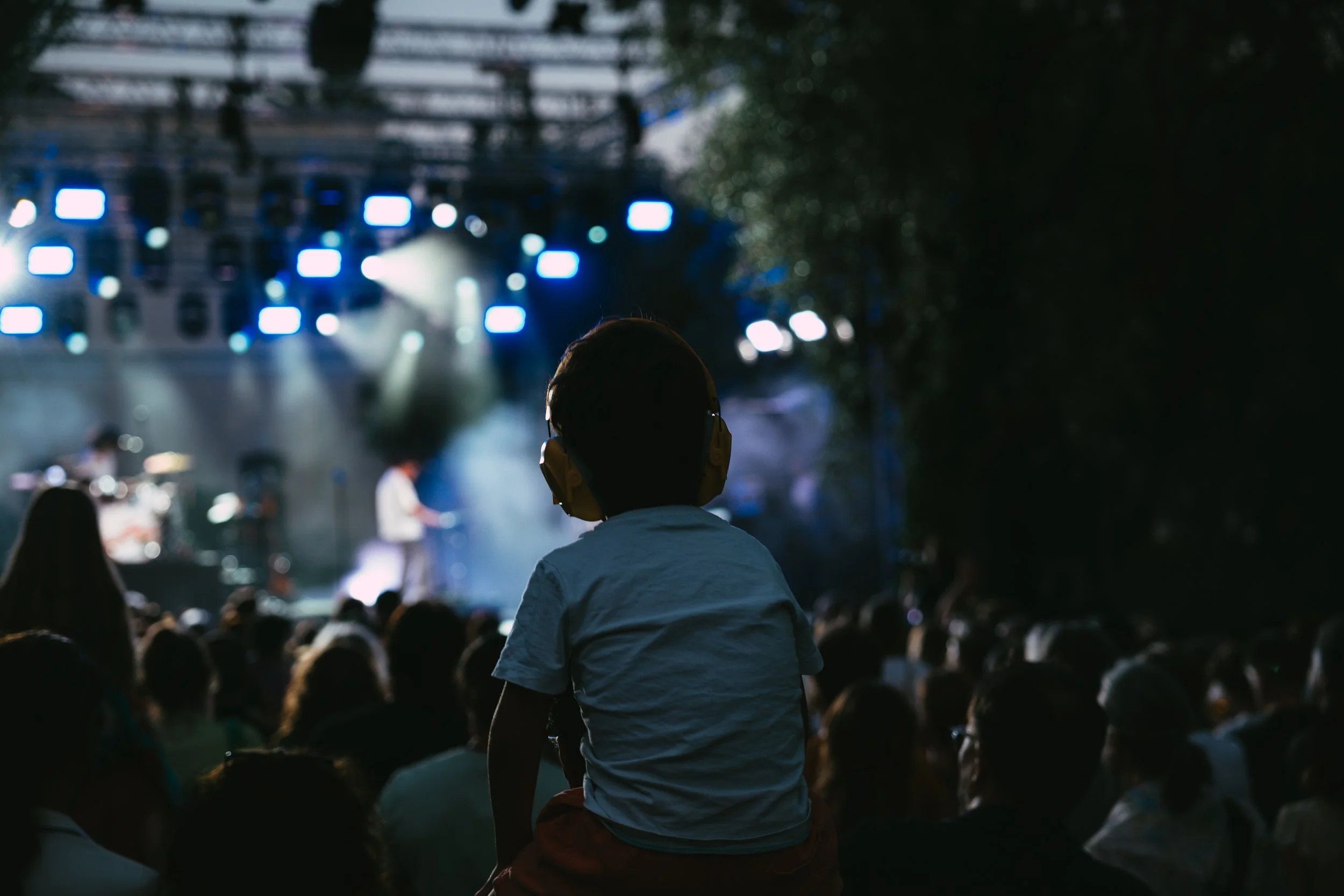 Person wearing headphones watching a concert or performance on a stage with blue lighting, with a crowd seated in front of the stage.