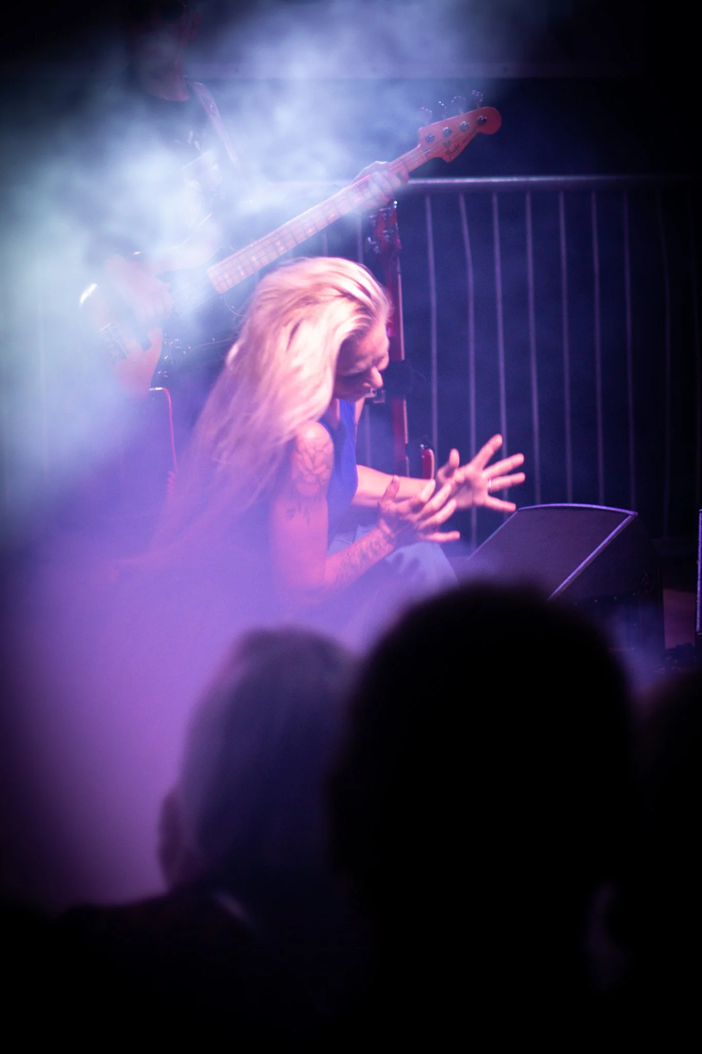 A woman performing guitar on stage with a bassist in background, surrounded by purple and blue stage lighting and fog. Audience silhouettes are visible at the bottom.