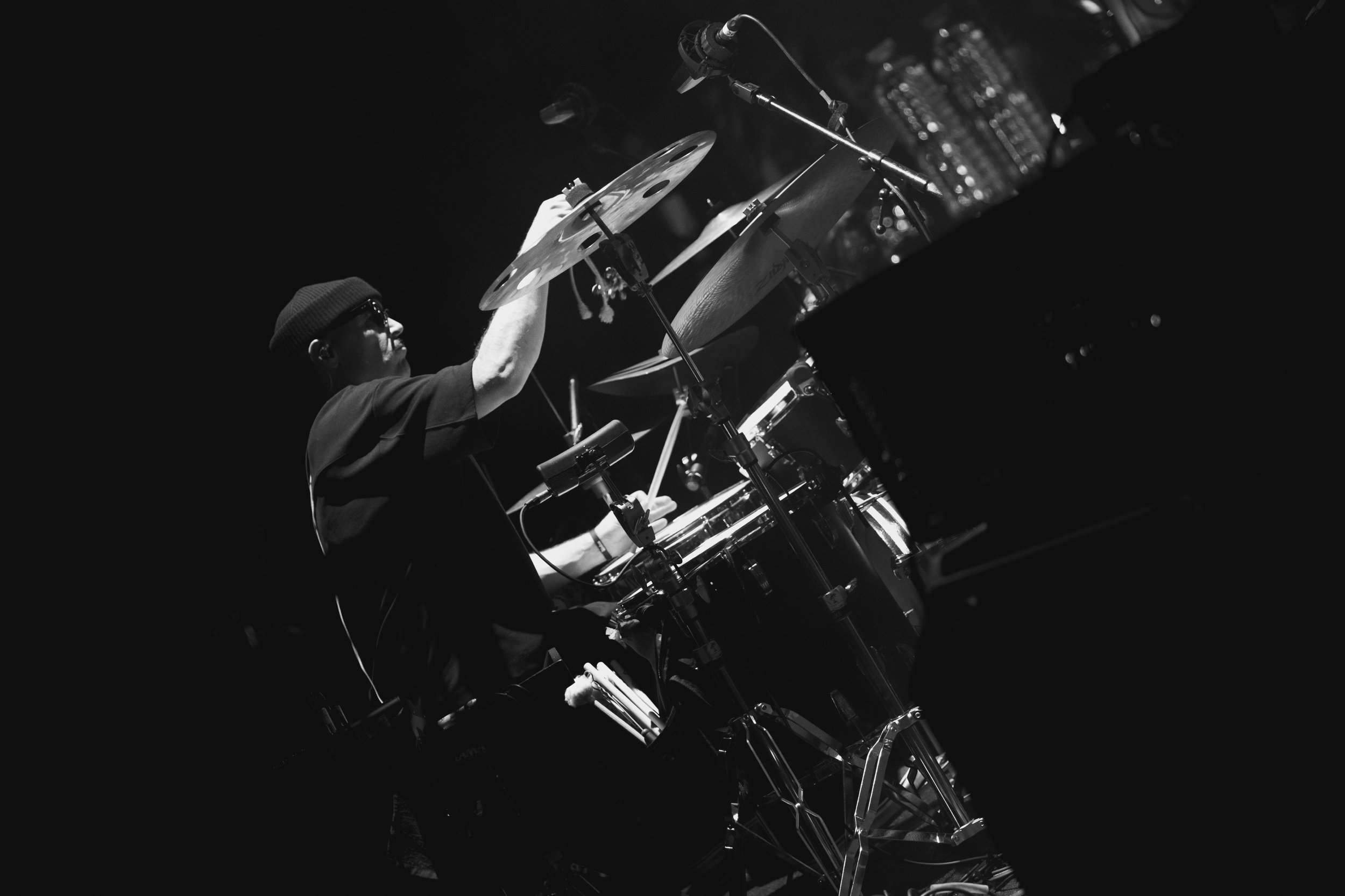 A black and white photo of a musician playing percussion instruments, wearing sunglasses and a beanie, with city lights in the background.