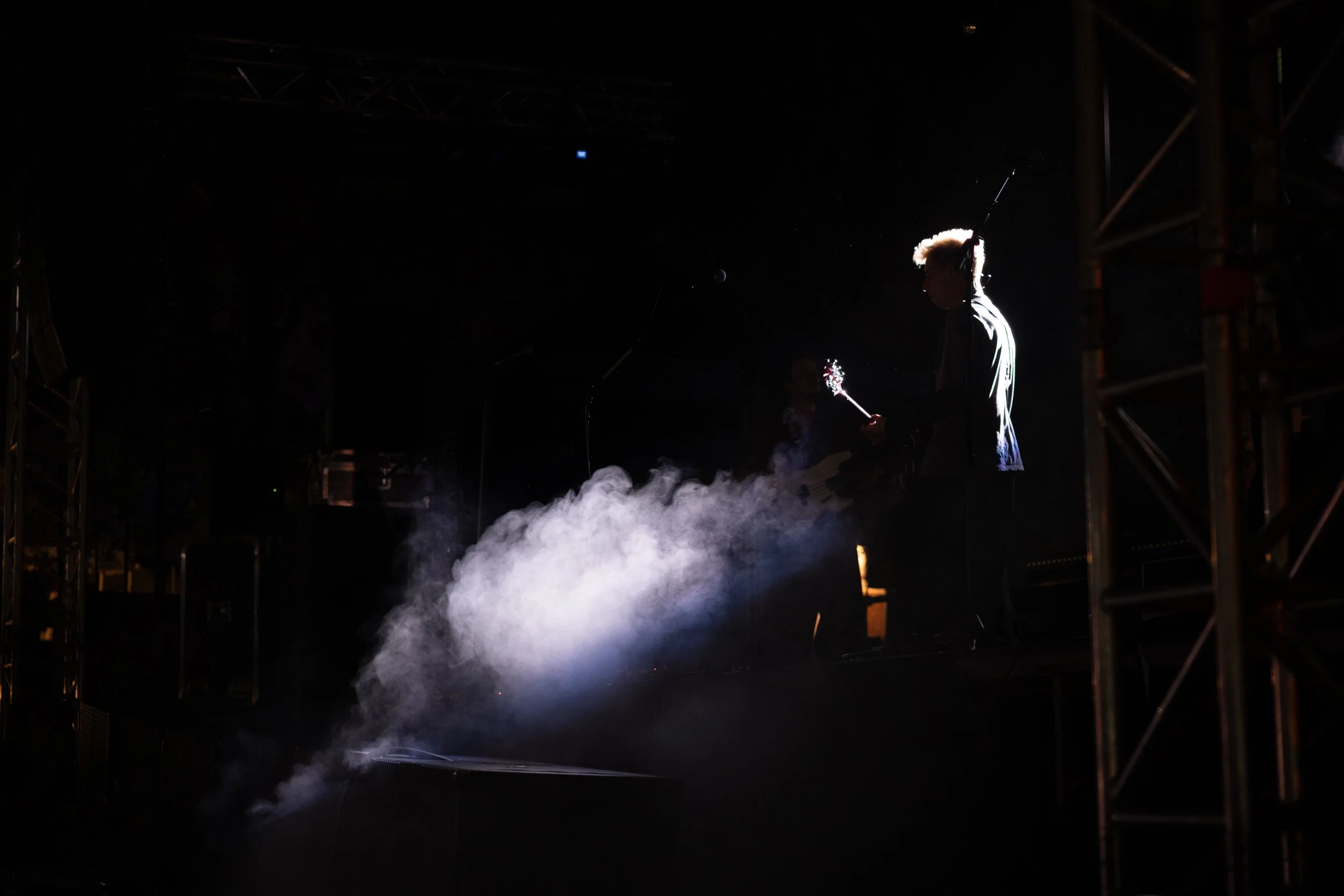 A musician performing on stage with a guitar, surrounded by smoke and backlit, with stage scaffolding visible on the sides.