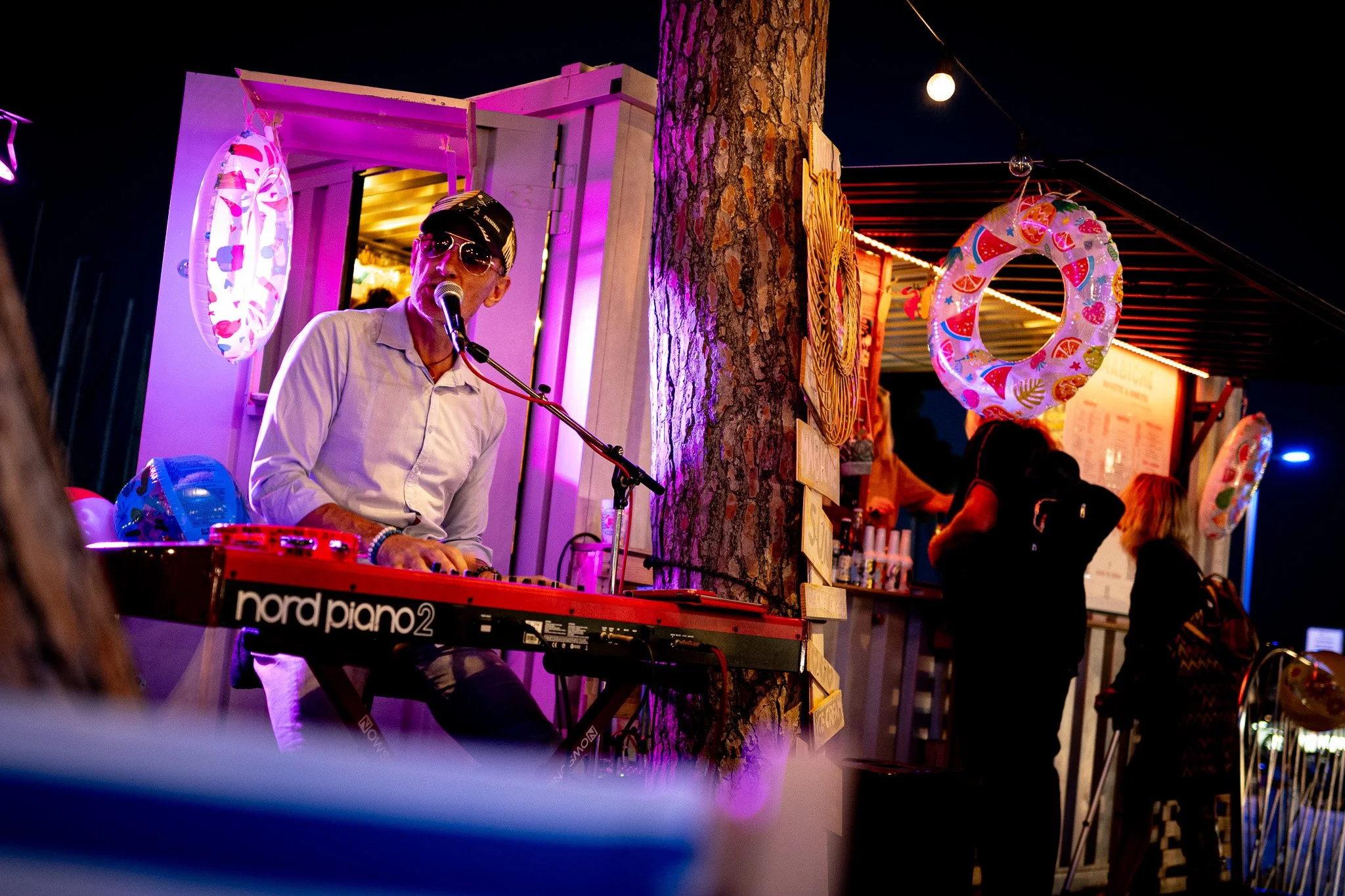 A man wearing sunglasses and a hat, playing a keyboard and singing into a microphone at an outdoor nighttime event. There are decorative ring-shaped inflatables hanging around, and a tree with wooden signs attached. People are seen in the background