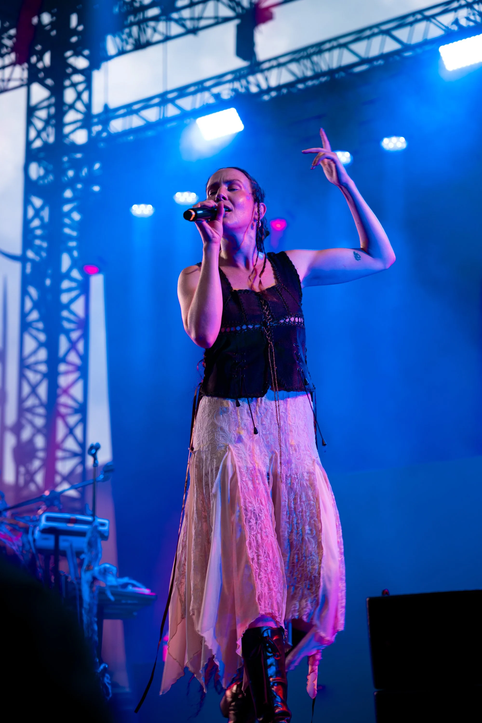 Female singer performing on stage with blue lighting, wearing a black top and long pink skirt, holding microphone, with stage equipment in the background.