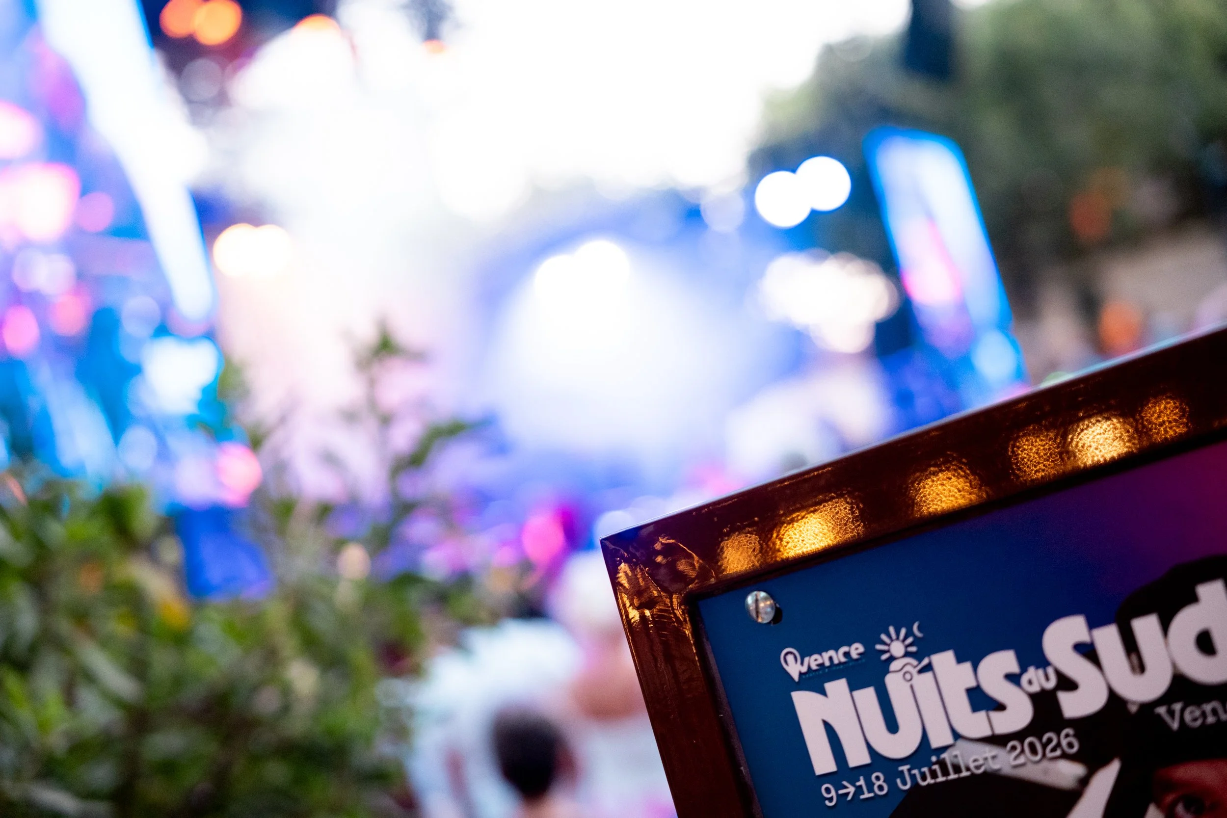 Blurred colorful lights with a partial view of a blue sign or poster in the foreground on the right side, possibly related to an event or festival.