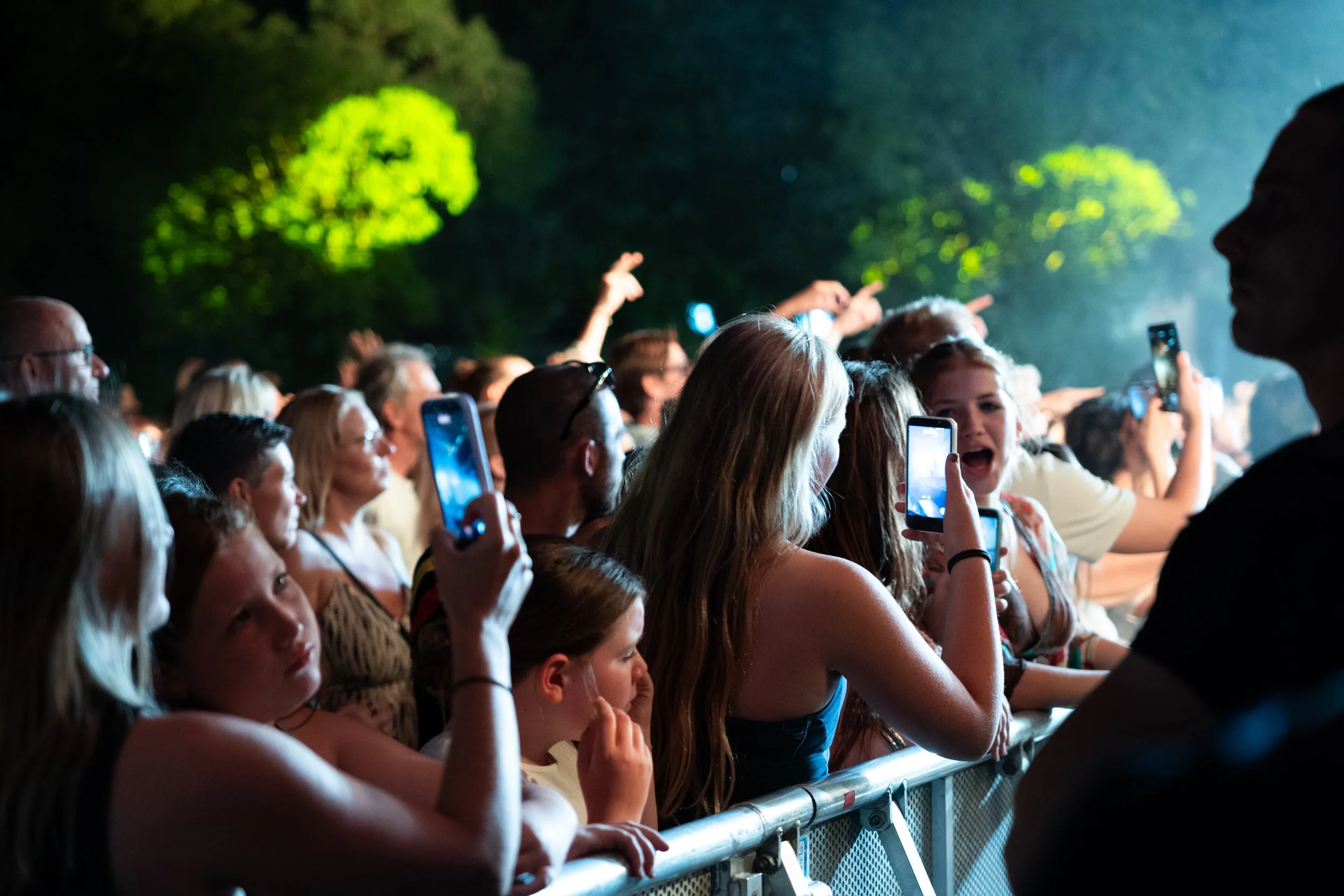 Crowd of people at an outdoor concert or event, some holding up phones to take pictures, with trees visible in the background at night.