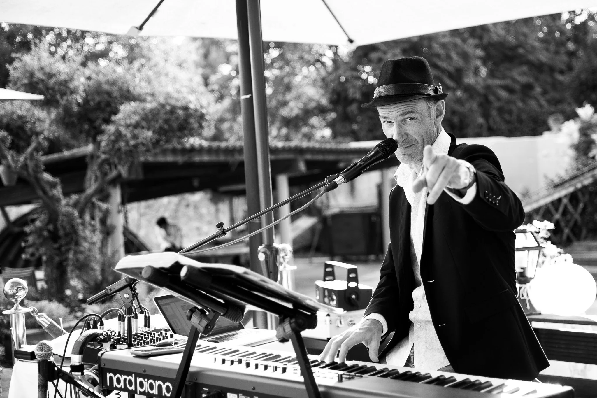 A man wearing a hat and suit points at the camera while playing a keyboard at an outdoor event.