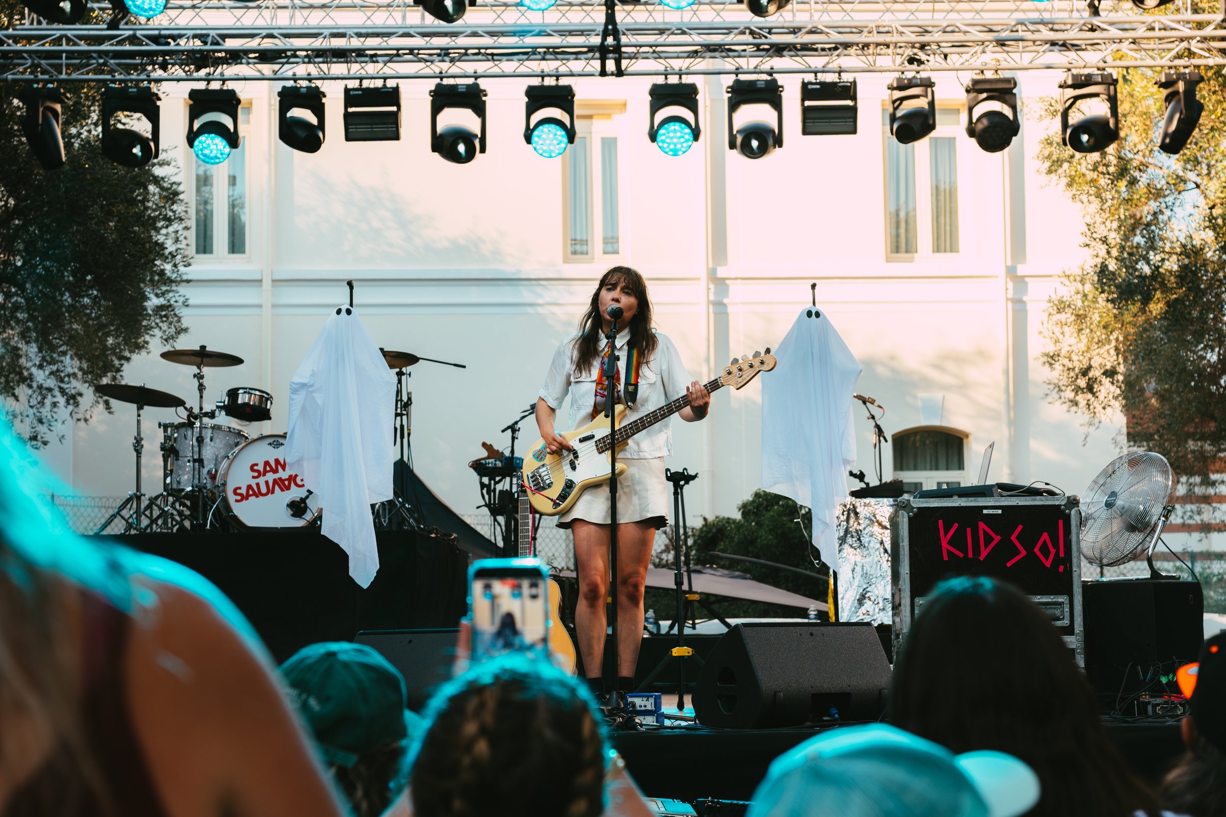 A female singer with long dark hair performs on stage holding a bass guitar, wearing a white outfit, at an outdoor concert with stage lights and musical instruments in the background.