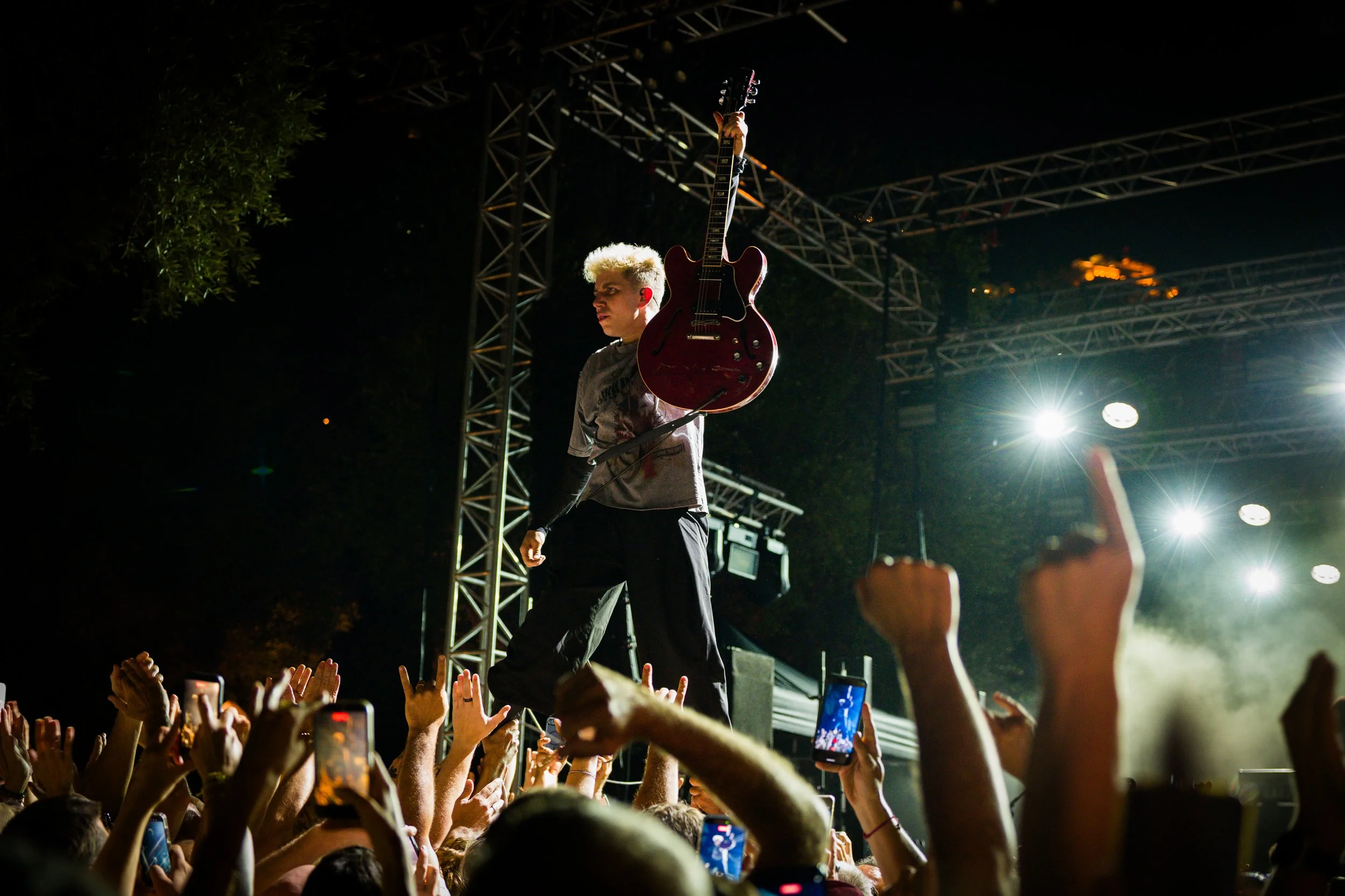 A musician with bleached blonde hair performing on stage at night, holding a guitar above his head, with a large crowd in front reaching towards him and many holding up phones