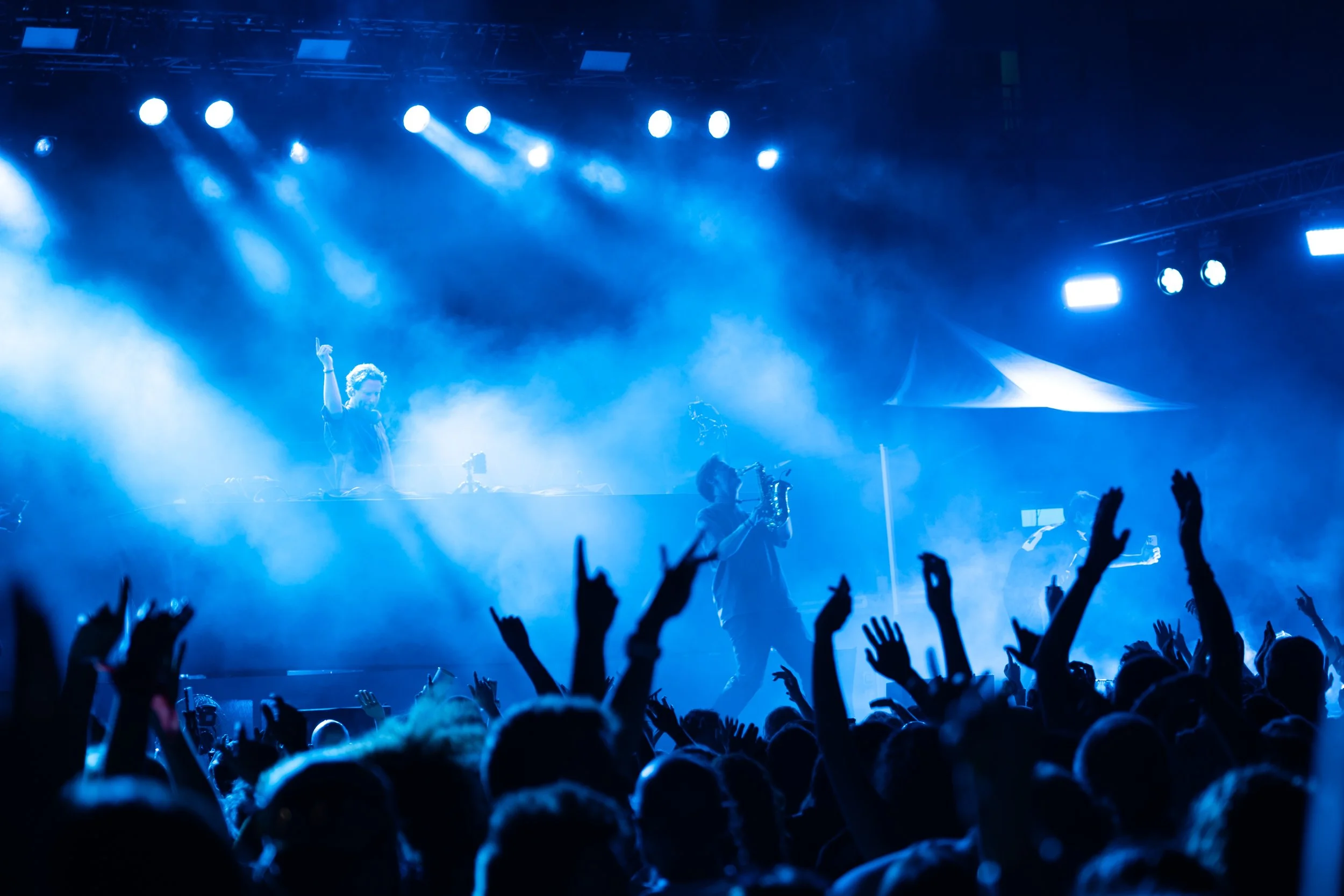 Concert scene with blue lighting, a DJ or performer on stage, and a crowd with raised hands.