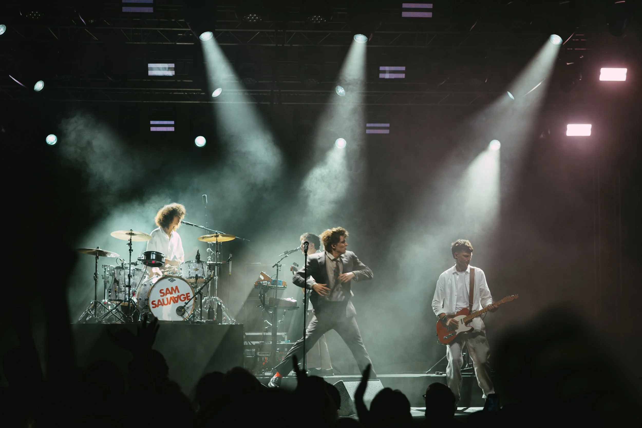 A band performing on stage with bright spotlights and smoke effects, with some audience members visible in the foreground.