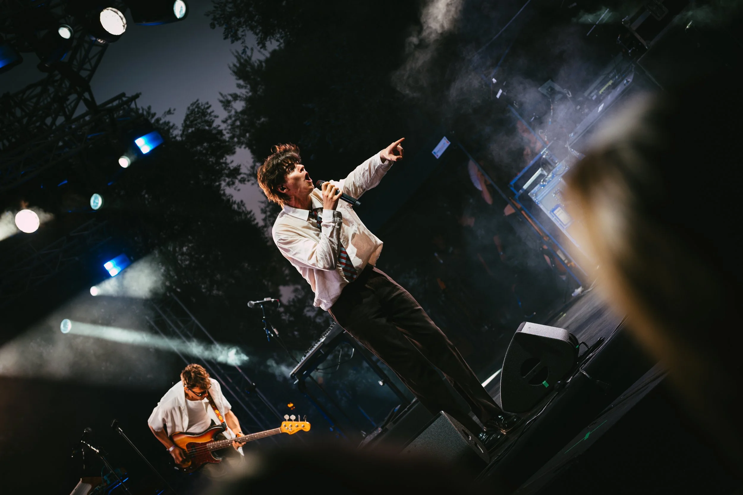 Musician in white shirt and striped tie performing with microphone at outdoor concert stage, guitarist in white shirt with sunglasses in foreground, stage lights and trees in background