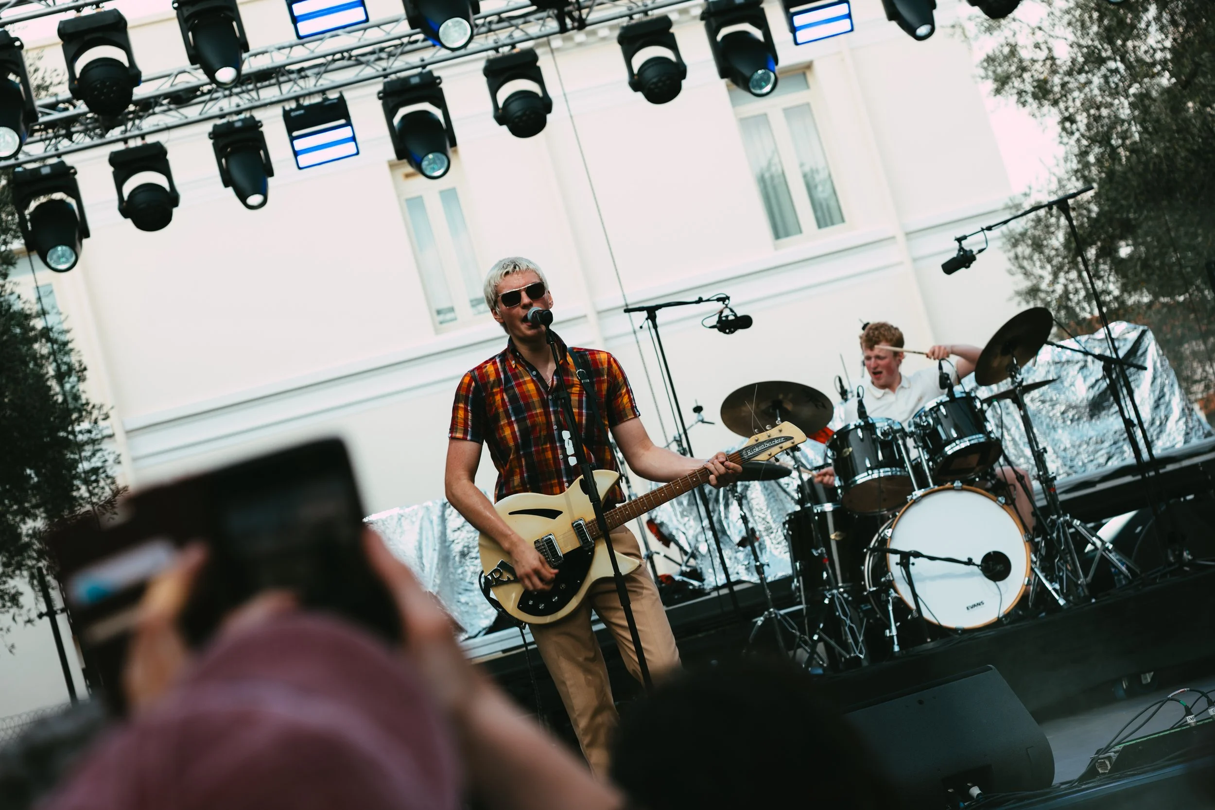 Musicians performing on an outdoor stage, with a guitarist singing into a microphone and a drummer playing in the background, under stage lights.
