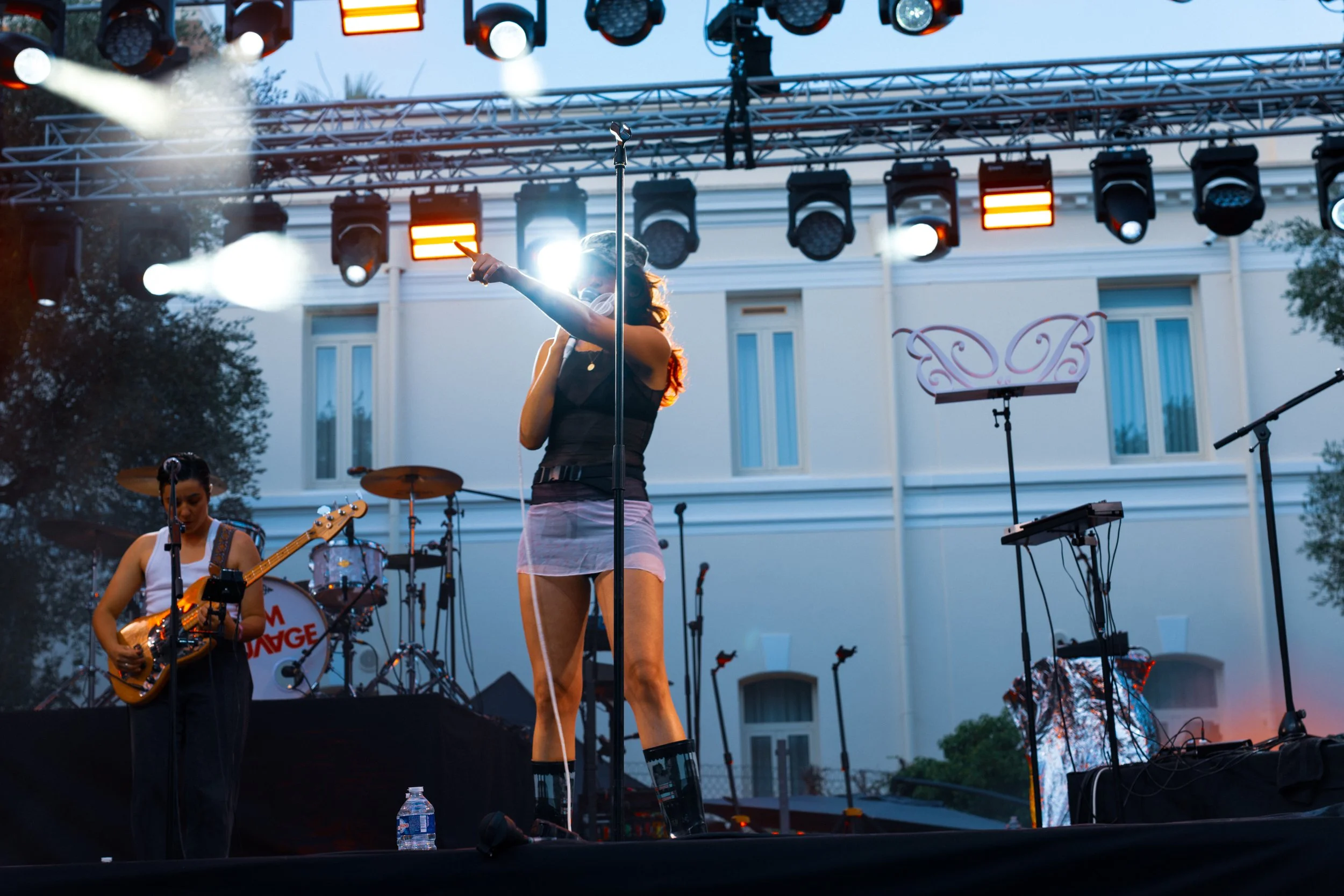 A female singer wearing a black top and pink shorts performs on an outdoor stage during twilight, with musicians and musical equipment around her and stage lighting overhead.