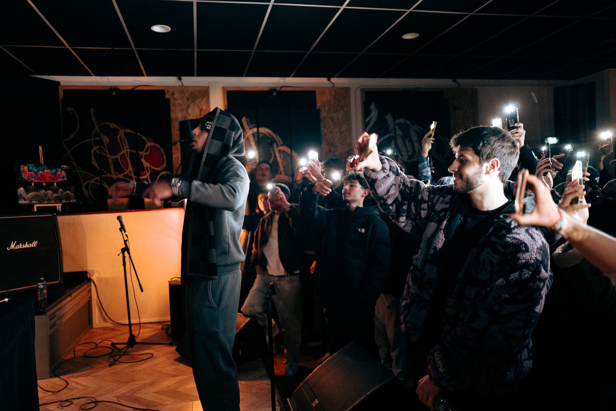 A performer on stage with a hoodie and scarf, facing away, at a concert venue with a crowd of young people taking photos with their phones.