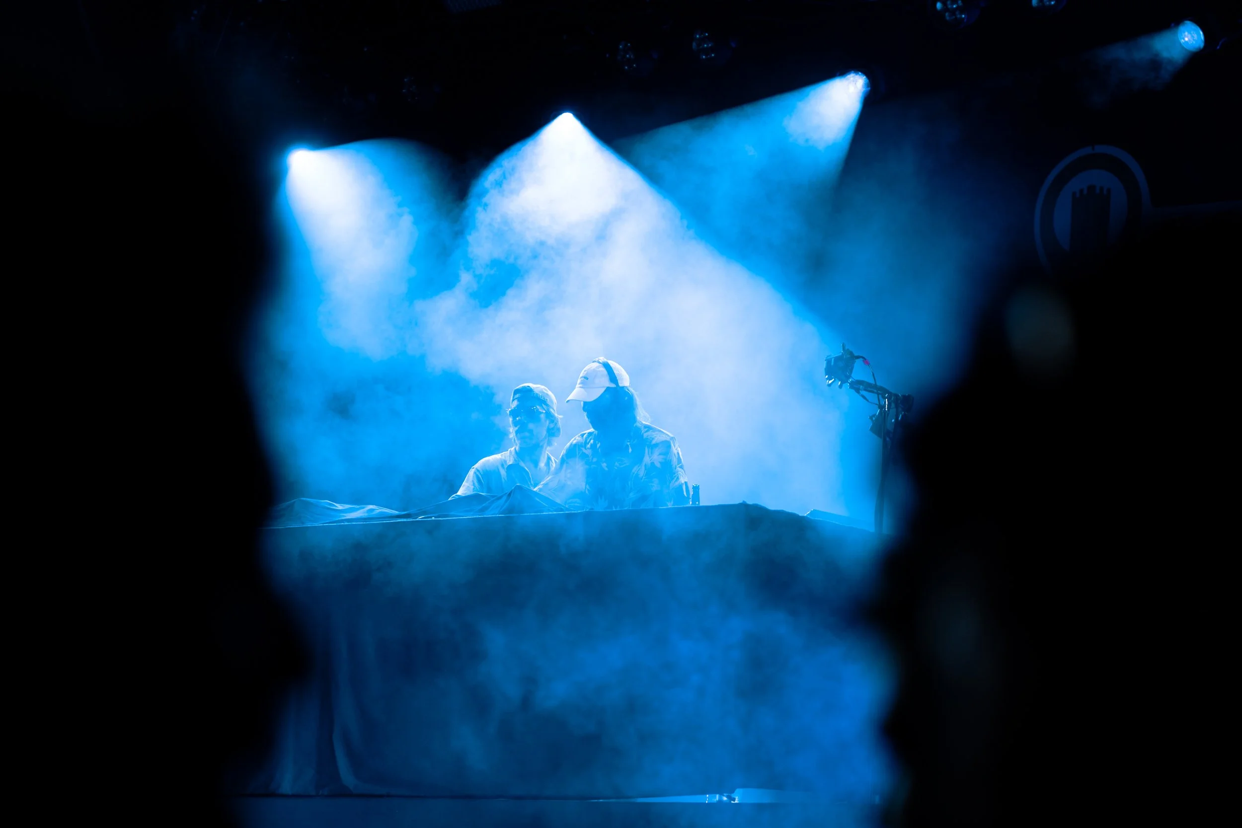 Two people wearing caps and glasses are sitting at a table and talking to each other, illuminated by blue stage lights during a performance or event.