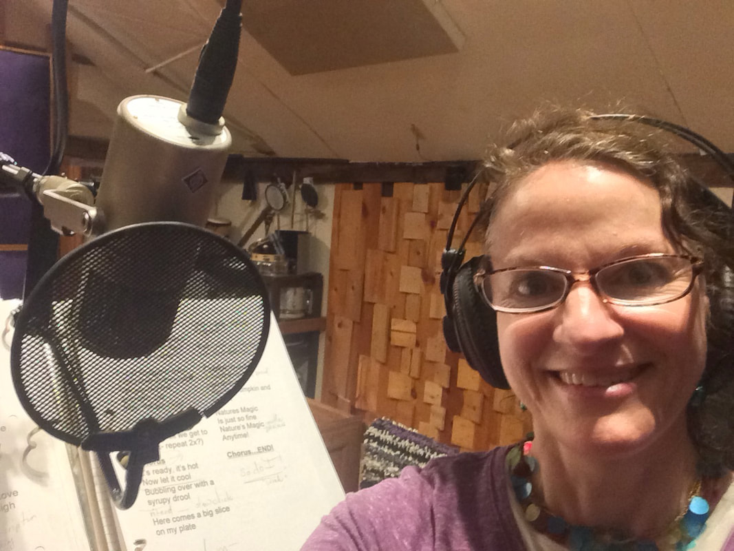 A woman with glasses and curly hair wearing headphones takes a selfie in a recording or music studio, with a large microphone and soundproofing panels in the background.