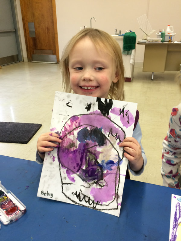 Young girl holding a colorful abstract painting of a face, smiling in an indoor setting with art supplies and a sink in the background.