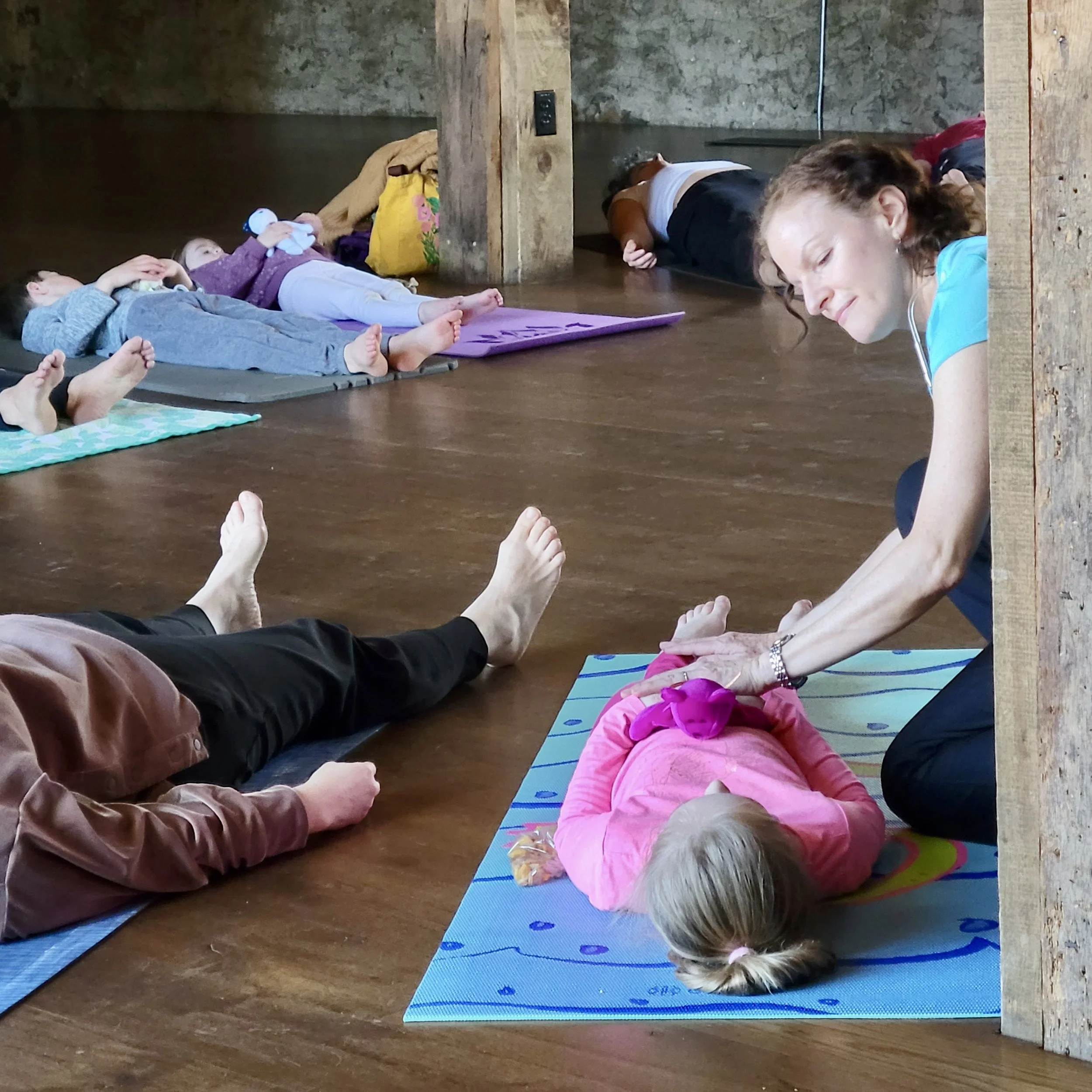 Children and adults lying on their stomachs on yoga mats, participating in a group relaxation or meditation session in a rustic room with wooden floors and a stone wall. The children are holding plush toys, and the adults are smiling and appear relax