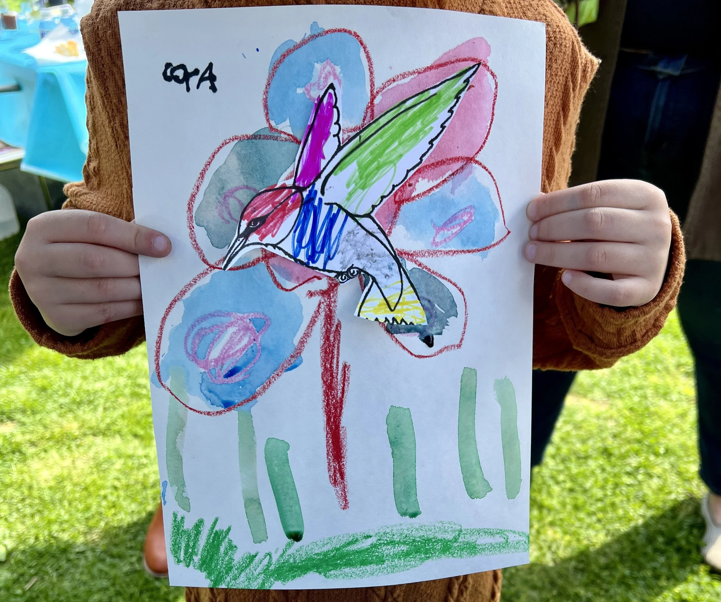 Child holding a brightly colored hand-drawn picture of a bird with a purple head, red body, blue wings, and green tail, against a background of blue and red flowers.