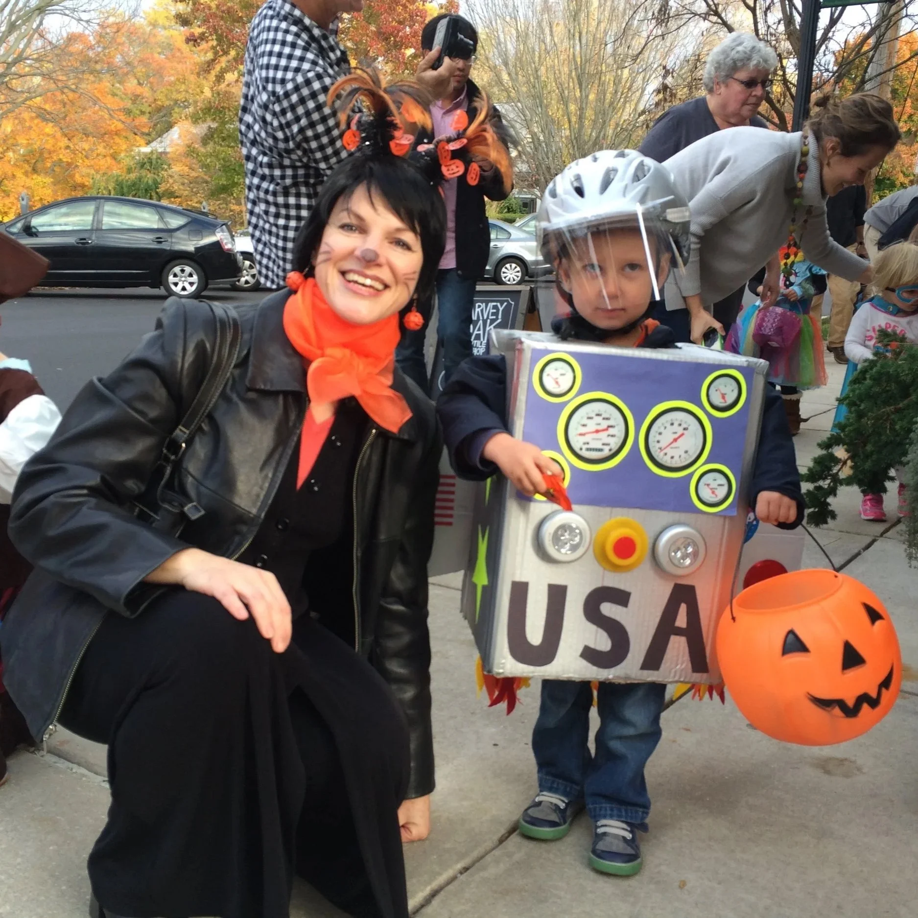 People dressed in Halloween costumes at an outdoor event during fall, including a woman with face paint and a young boy with a helmet and costume, and a woman in a white outfit. There are decorated pumpkins and a sign that reads 'Survey' in the backg