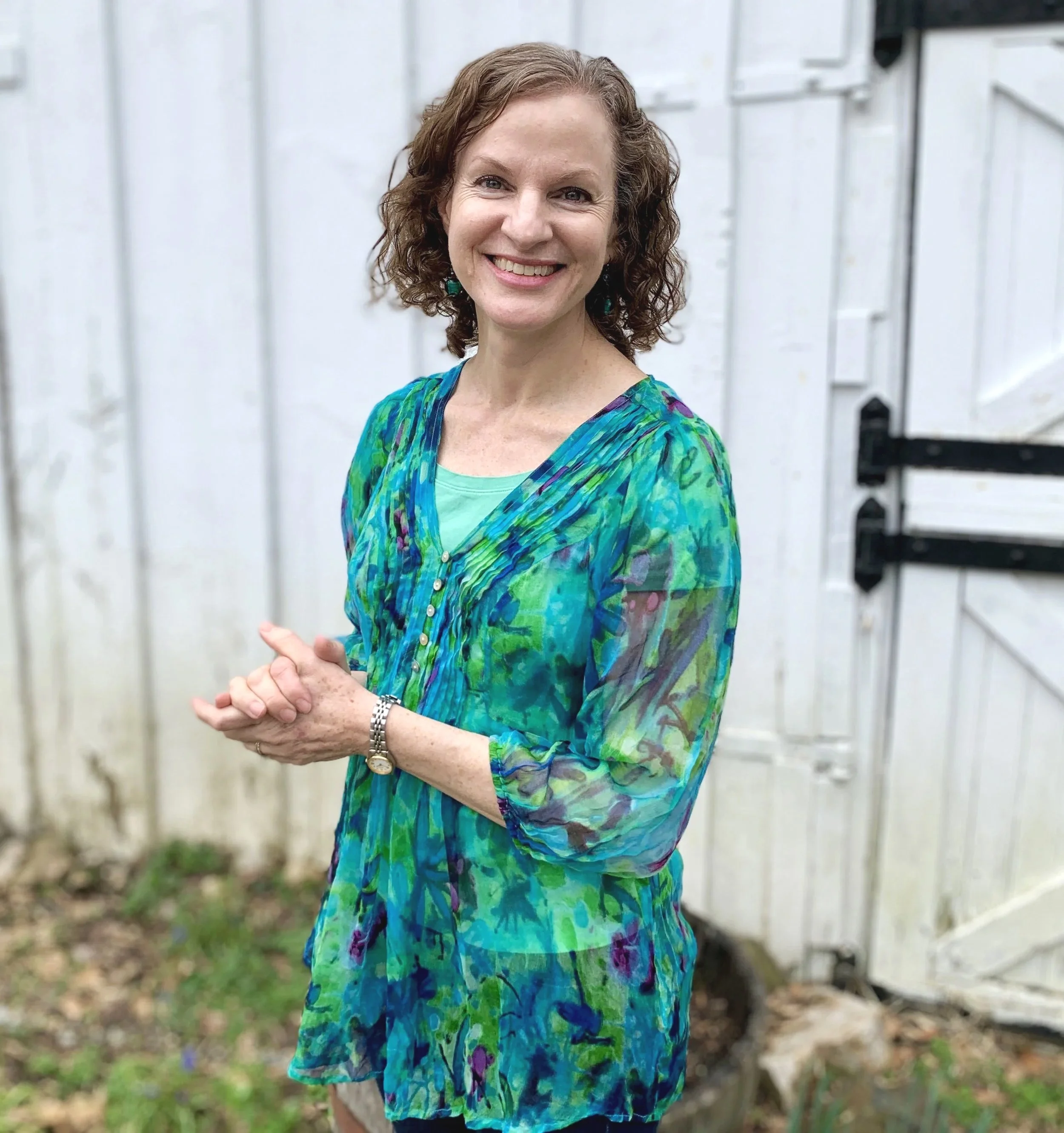 Smiling woman with curly hair in a colorful dress outdoors in front of a white wooden fence.