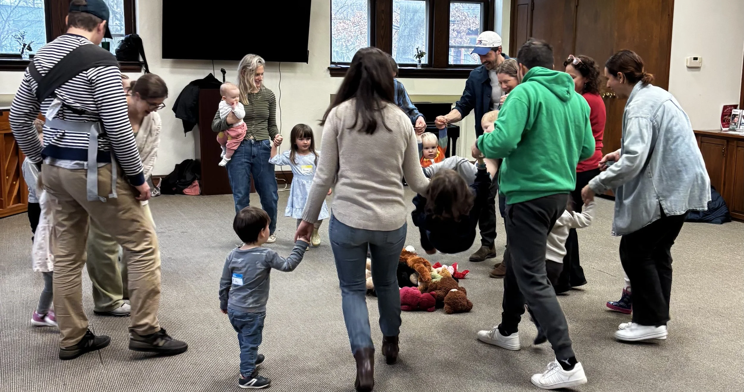 Group of children and adults holding hands in a circle playing a game indoors, with stuffed animals on the floor in the center.