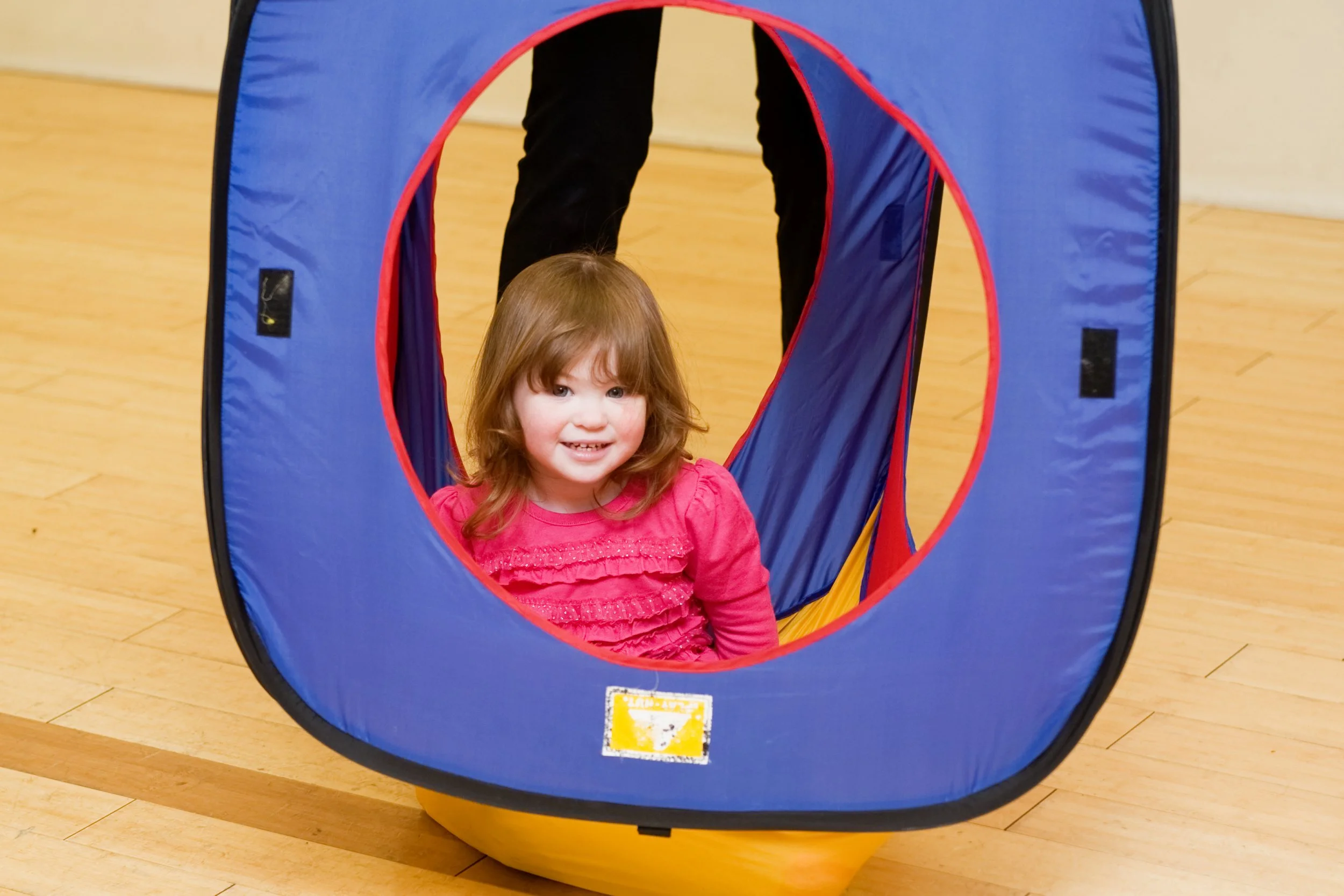 A young girl with brown hair and a pink dress sitting inside a blue and red play tunnel on a wooden floor.