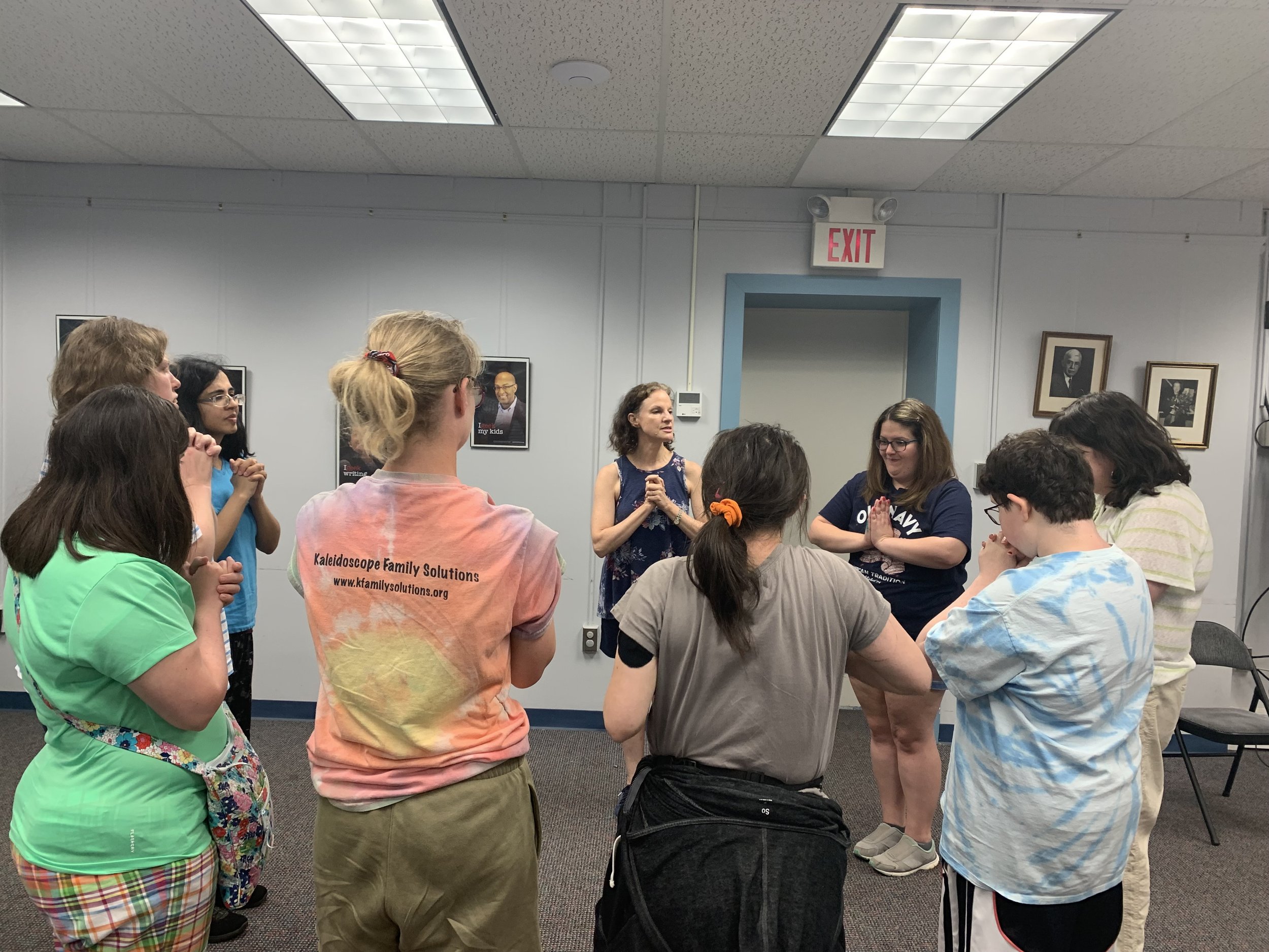 A group of neurodiverse young women people in a circle in an indoor room, participating in a group yoga activity  with Allison, with some people having their hands clasped in standing prayer pose.